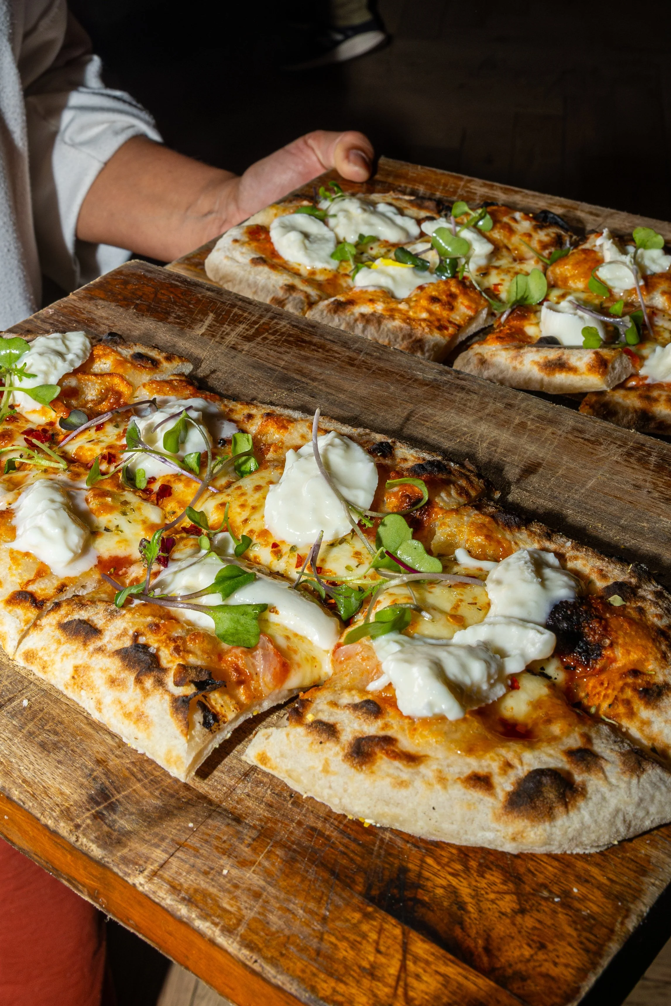 Close-up of a rectangular pizza with cheese, dollops of ricotta cheese, and microgreens on a wooden serving board.