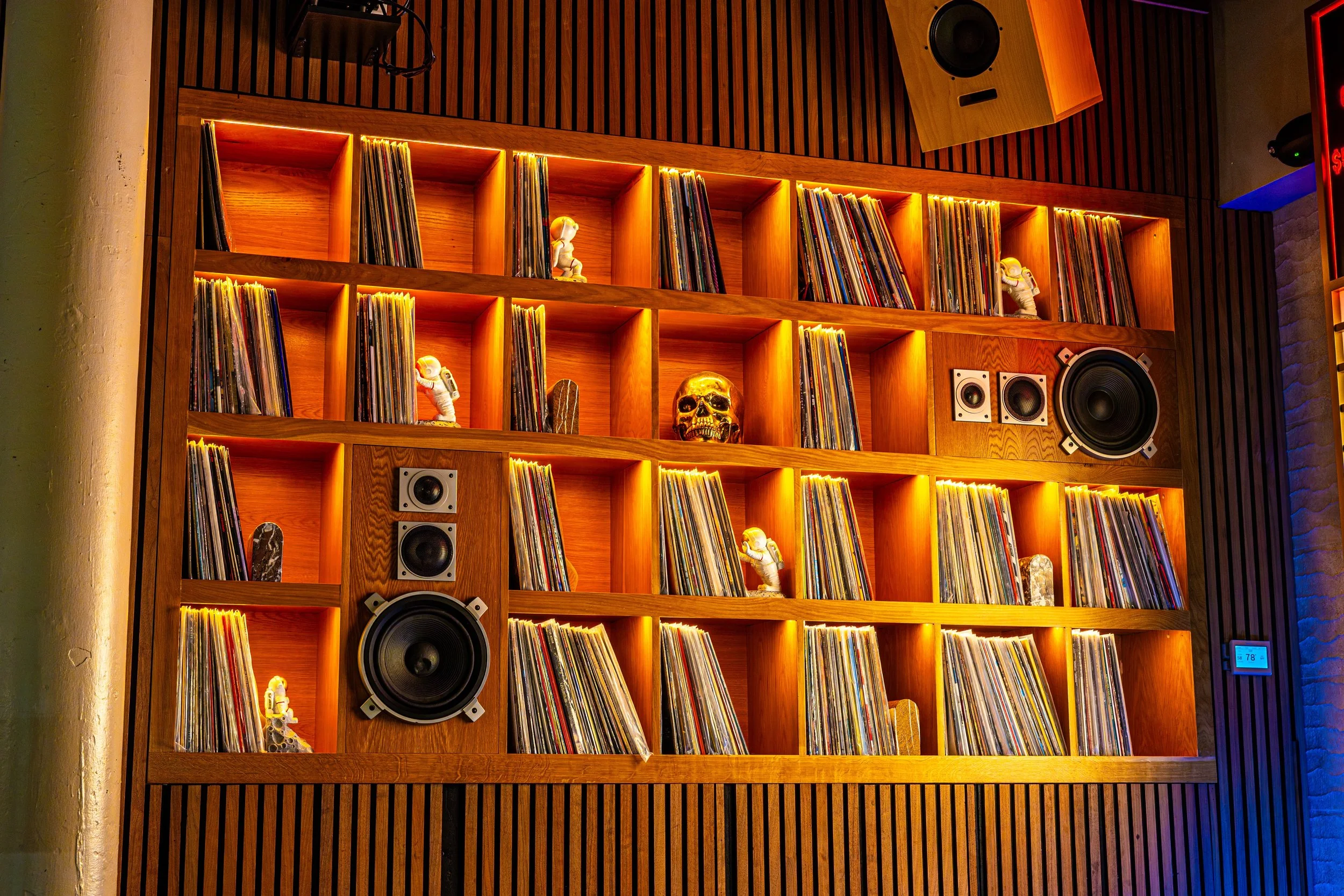 A wooden shelves filled with vinyl records, decorated with small astronaut figurines and a skull, and equipped with speakers. The scene is warmly lit with yellow and orange lighting.