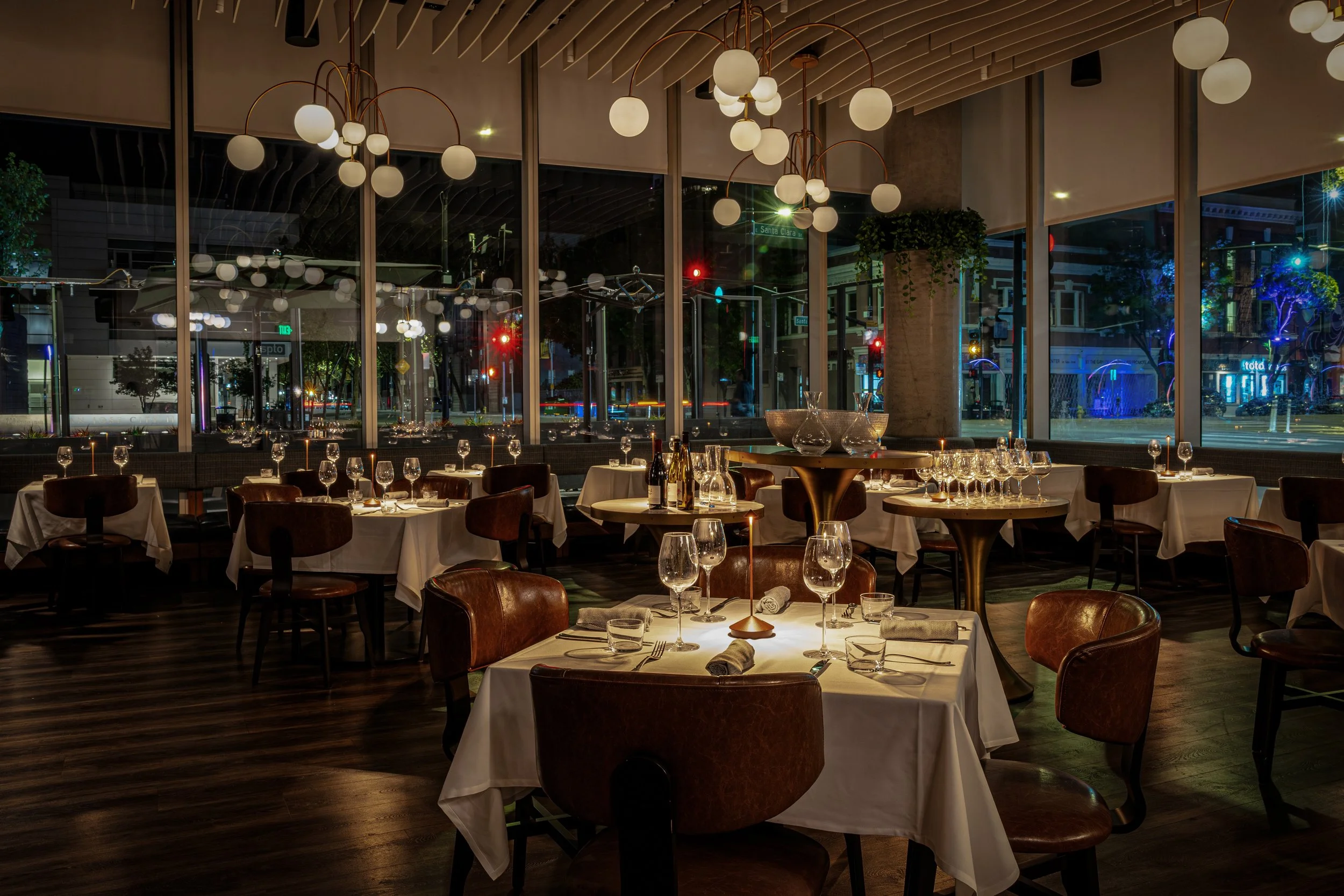 Interior of an upscale restaurant with large windows showing city street lights outside, decorated with modern hanging lamps, set tables with wine glasses, plates, and cutlery, and wooden flooring.