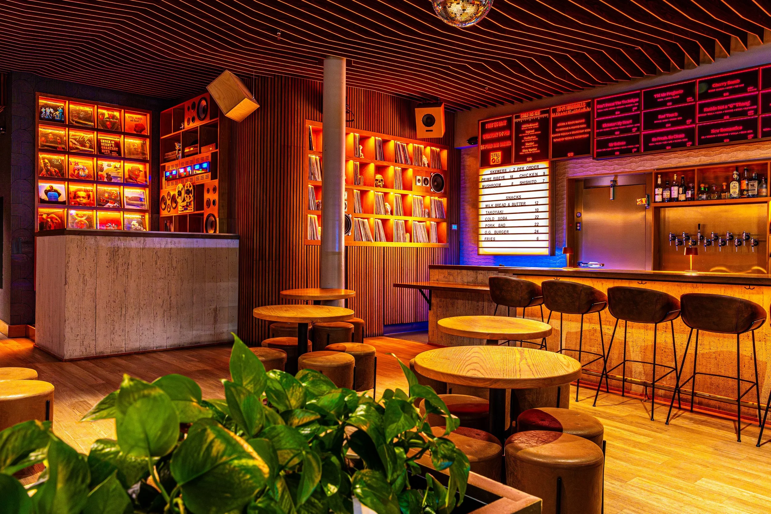 Interior of a stylish bar or lounge with wooden furniture, potted plants, vinyl records on display, and a brightly lit menu board.