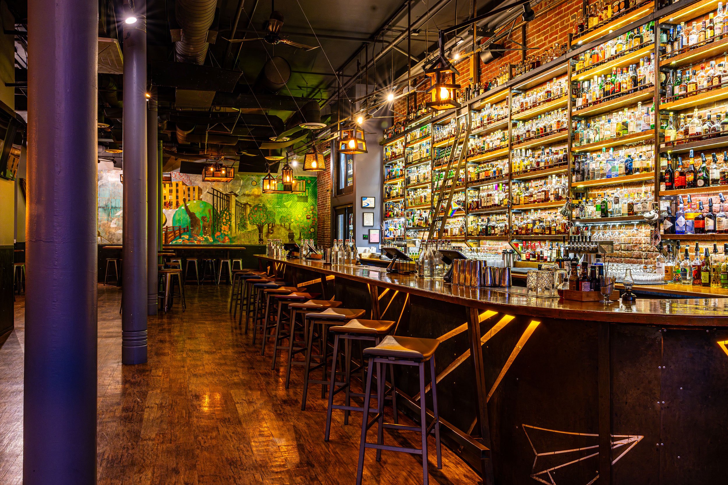 Interior of a bar with shelves of various liquor bottles, a long wooden counter with barstools, and colorful wall art in the background.