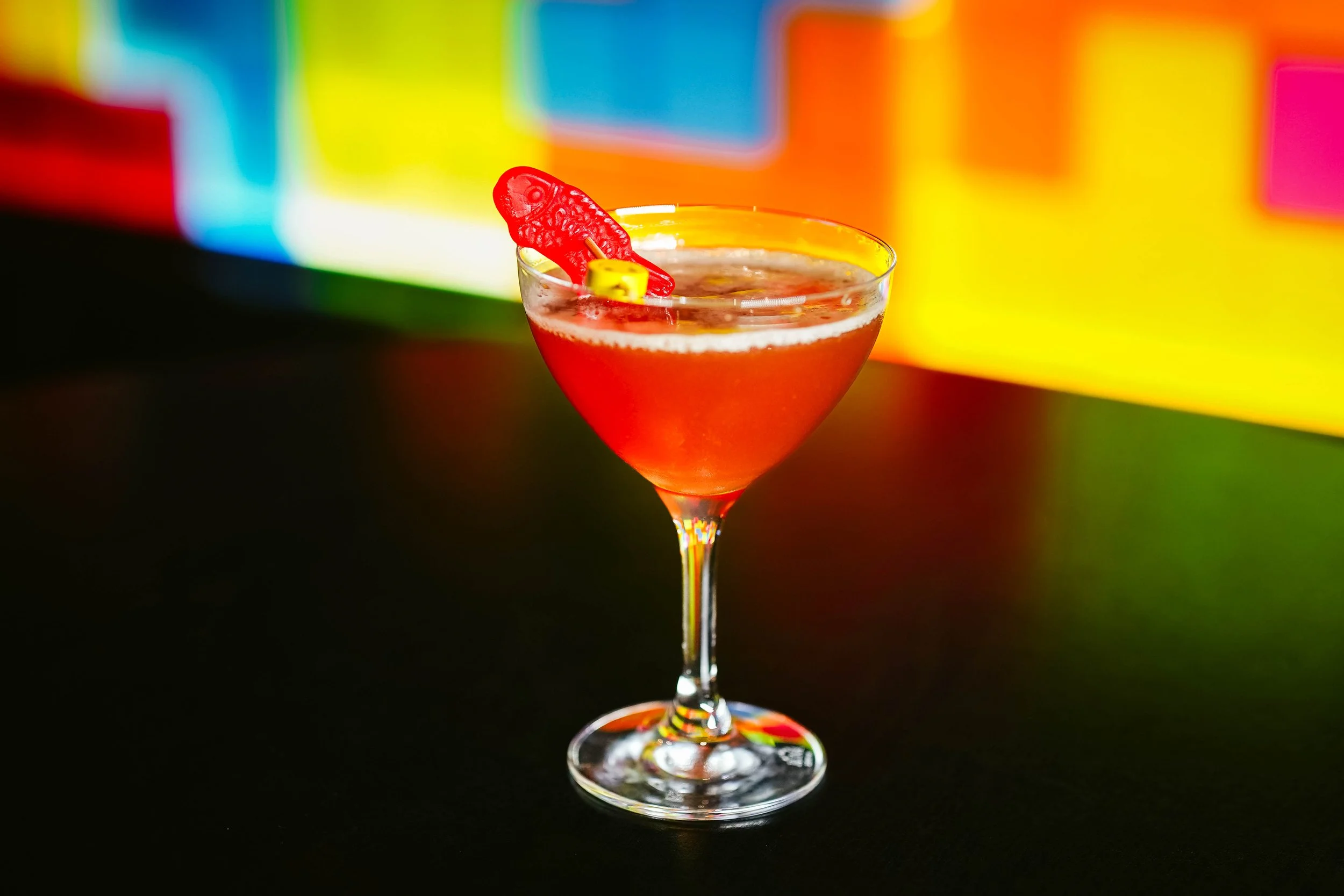 A colorful cocktail in a martini glass with a red fish-shaped gummy candy garnish, set against a blurred vibrant, multicolored background.