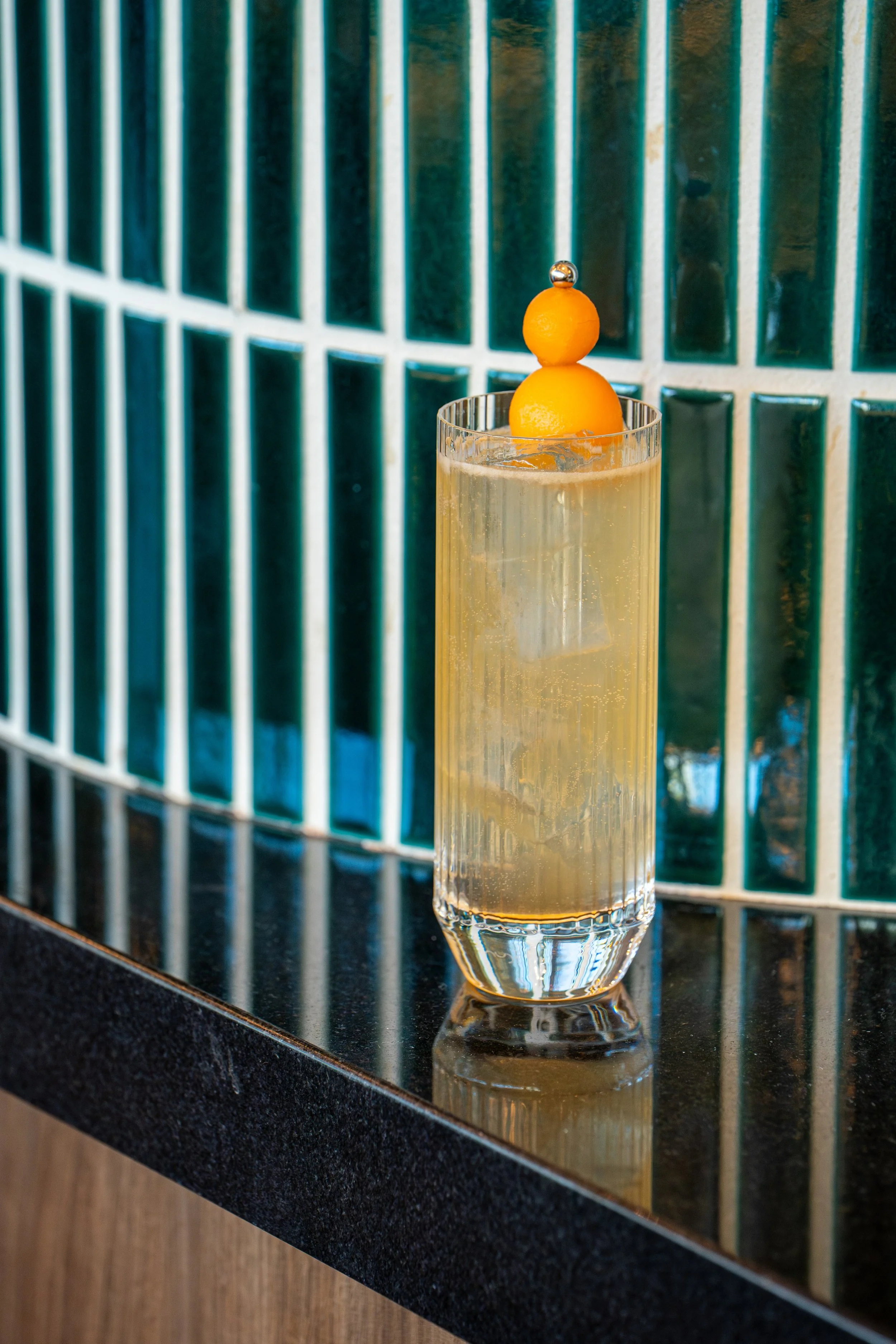 A tall clear glass filled with a light-colored beverage, garnished with two orange melon balls and a small metal ball on top, placed on a dark countertop reflecting the glass, with a background of green and white tiled wall.