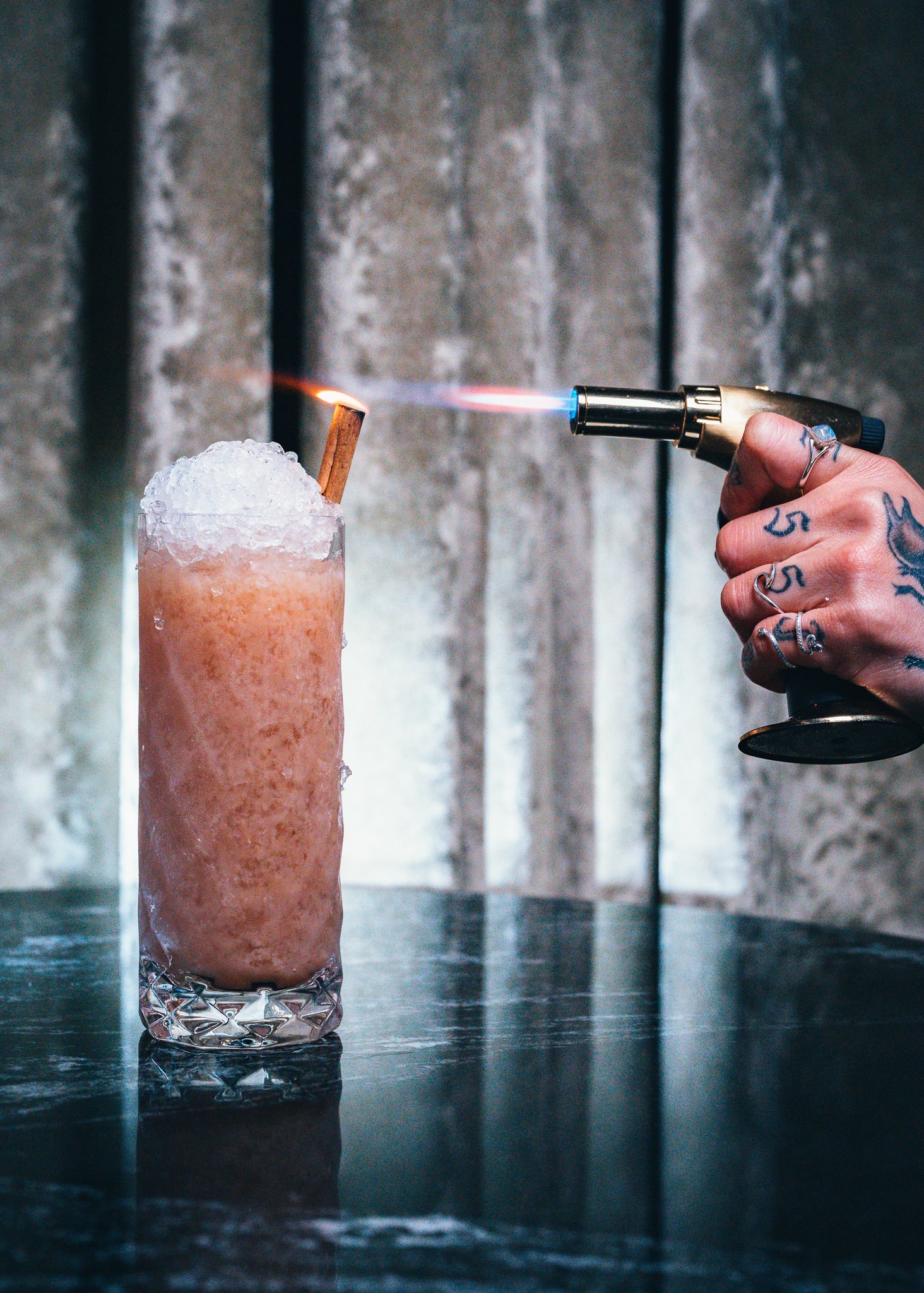 A hand holding a flamethrower aimed at a tall glass of pink frozen drink with ice crystals, garnished with a cinnamon stick. The drink is on a reflective dark surface.