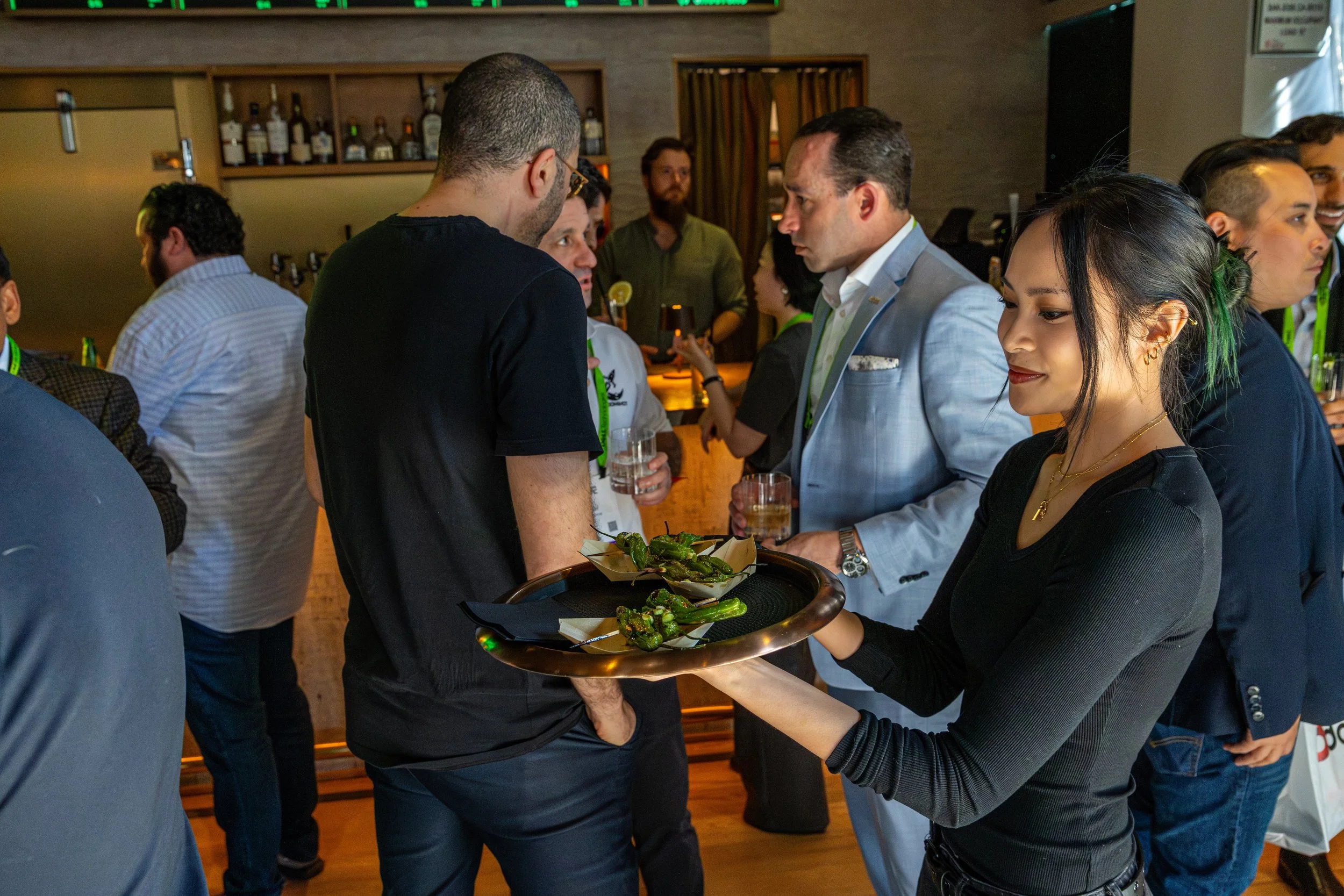 A woman is serving food to a man at a social gathering or event, with a tray of grilled green peppers in her hands. Other people are visible in the background, engaged in conversation.