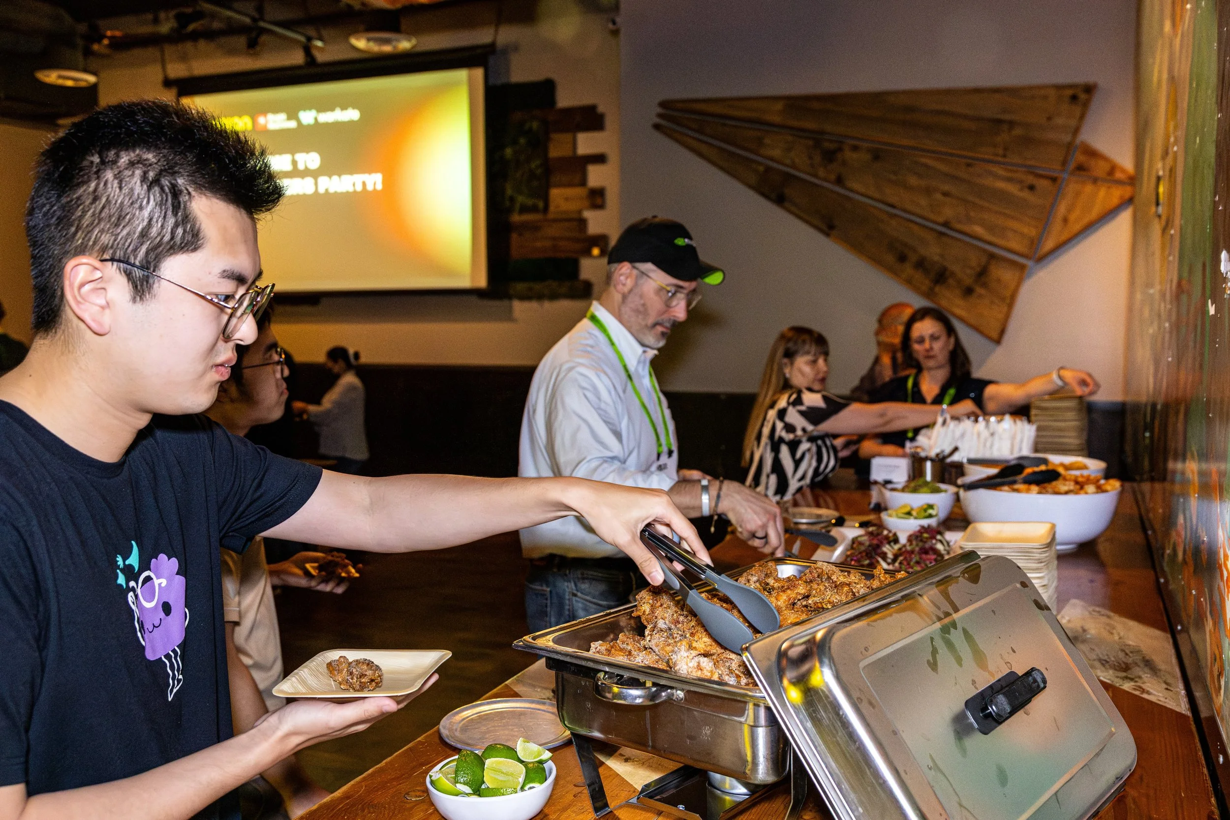 People serving themselves food from a buffet at a party, with bowls of various dishes and lime wedges on the counter.