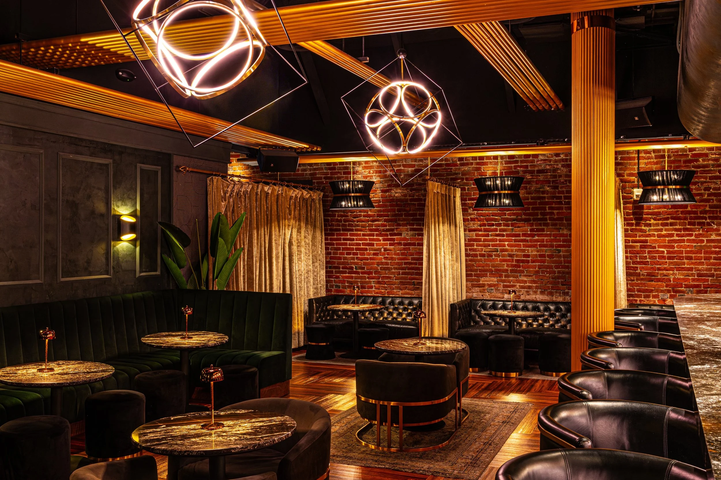 Interior of a modern lounge or upscale bar with hanging geometric light fixtures, brick wall, curtains, dining tables, and black leather chairs.