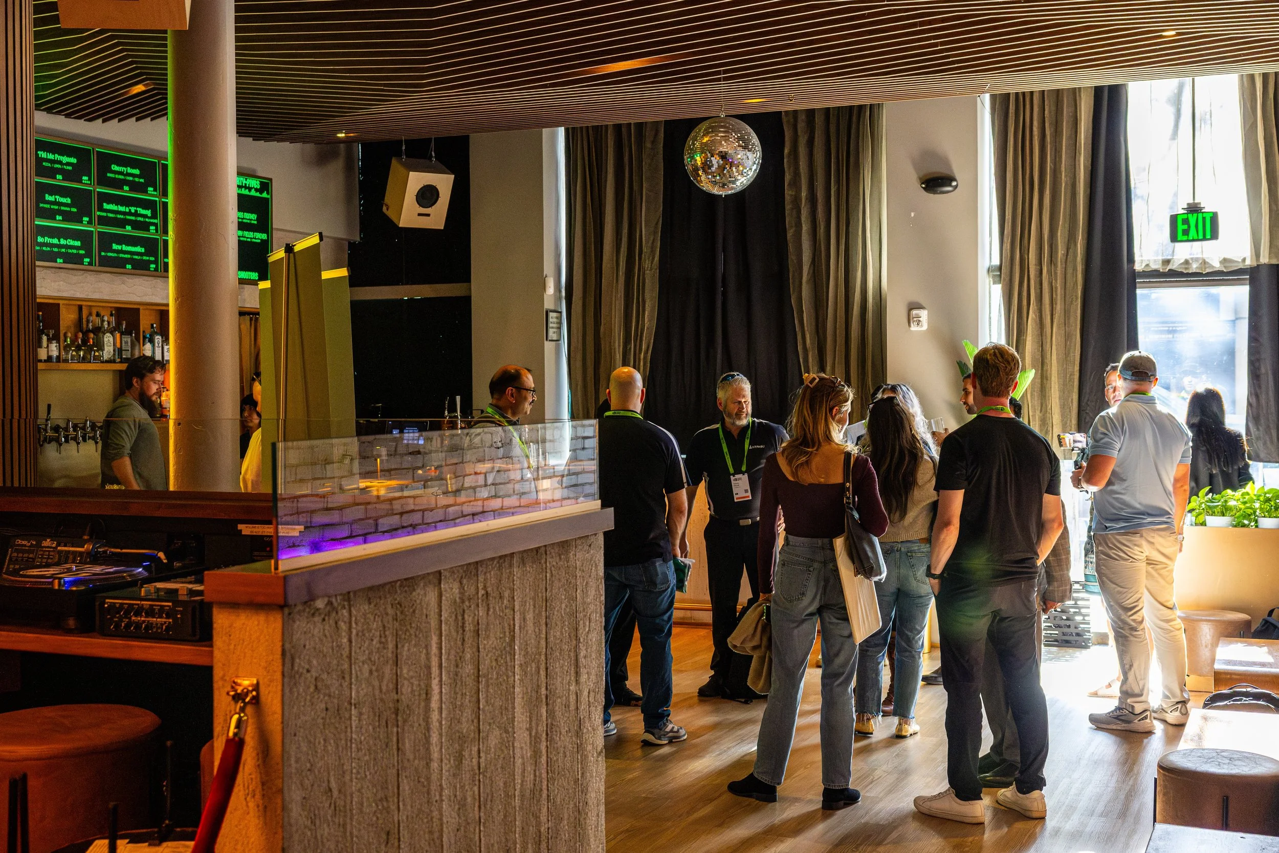 Guests gathered inside a modern bar or restaurant, with some standing near the bar counter, chatting, and others near large windows with curtains. There is a disco ball hanging from the ceiling, large green exit sign, and digital menu boards behind the bar. The atmosphere appears lively and casual.