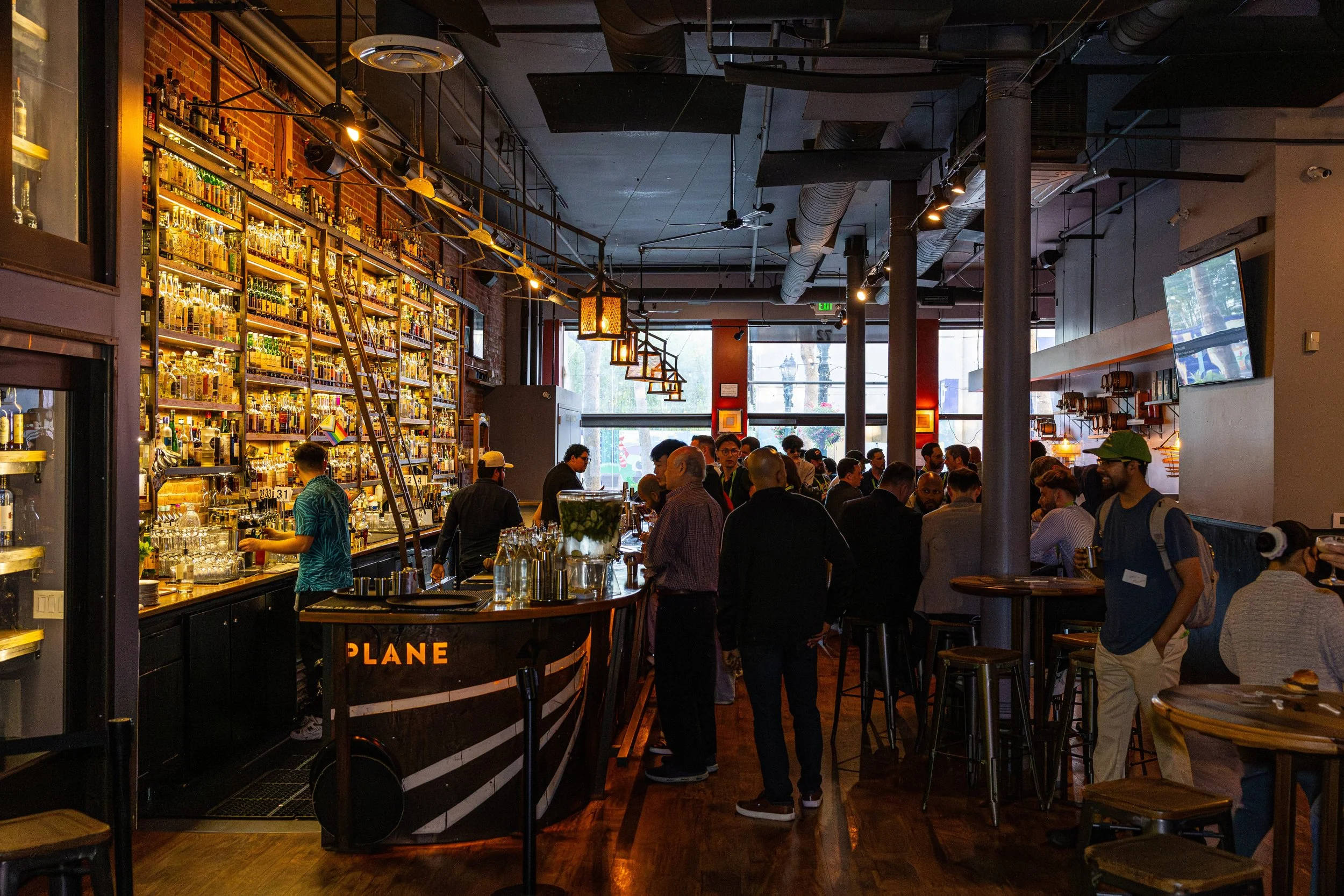 Crowded bar with people socializing, shelves stocked with liquor bottles, and a bartender preparing drinks.