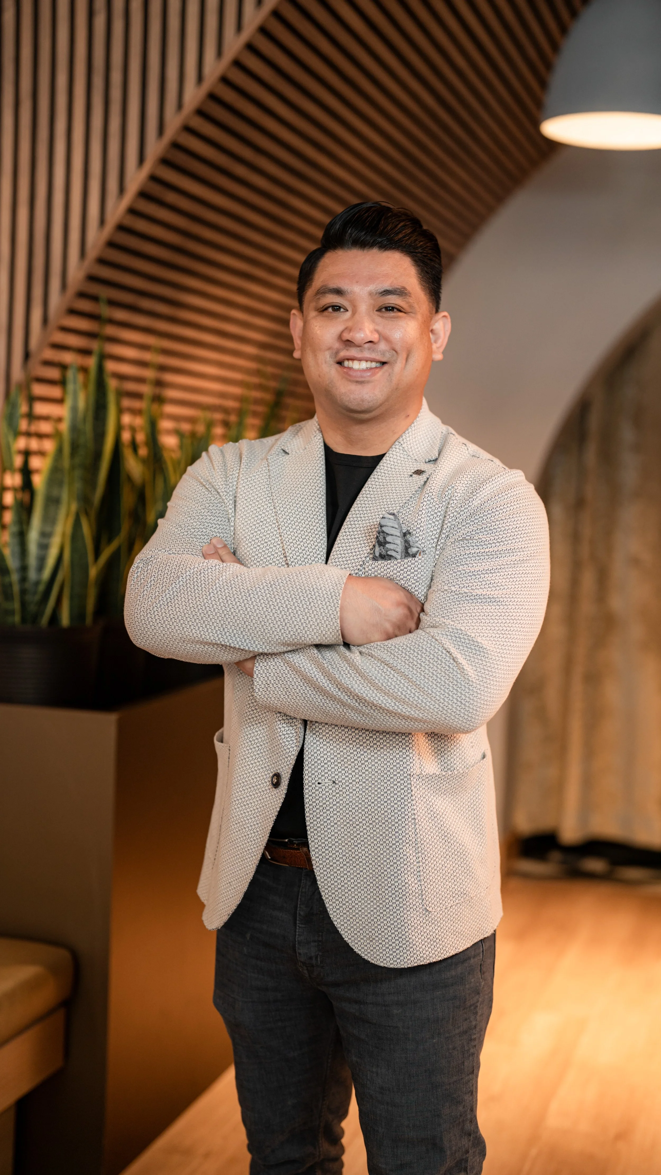 A man in a light-colored blazer with a pocket square, black shirt, and dark jeans, standing with arms crossed in an indoor setting with wooden accents and plants.