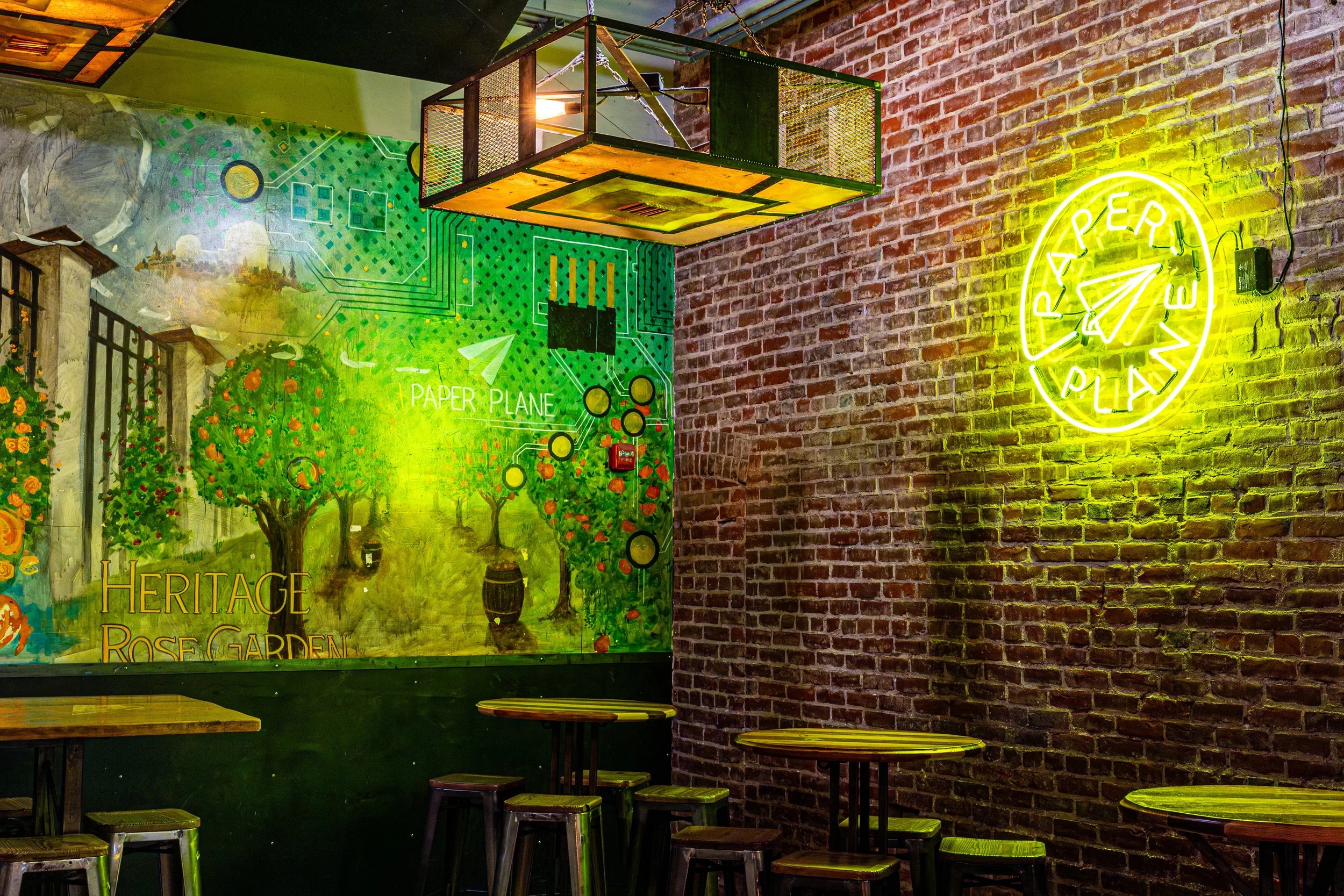 Interior of a restaurant or cafe with a brick wall, a colorful mural of a rose garden, and a neon sign that reads 'Paper Plane.' There are small round tables with stools against the wall. A ceiling-mounted structure made of chain-link fencing is visi