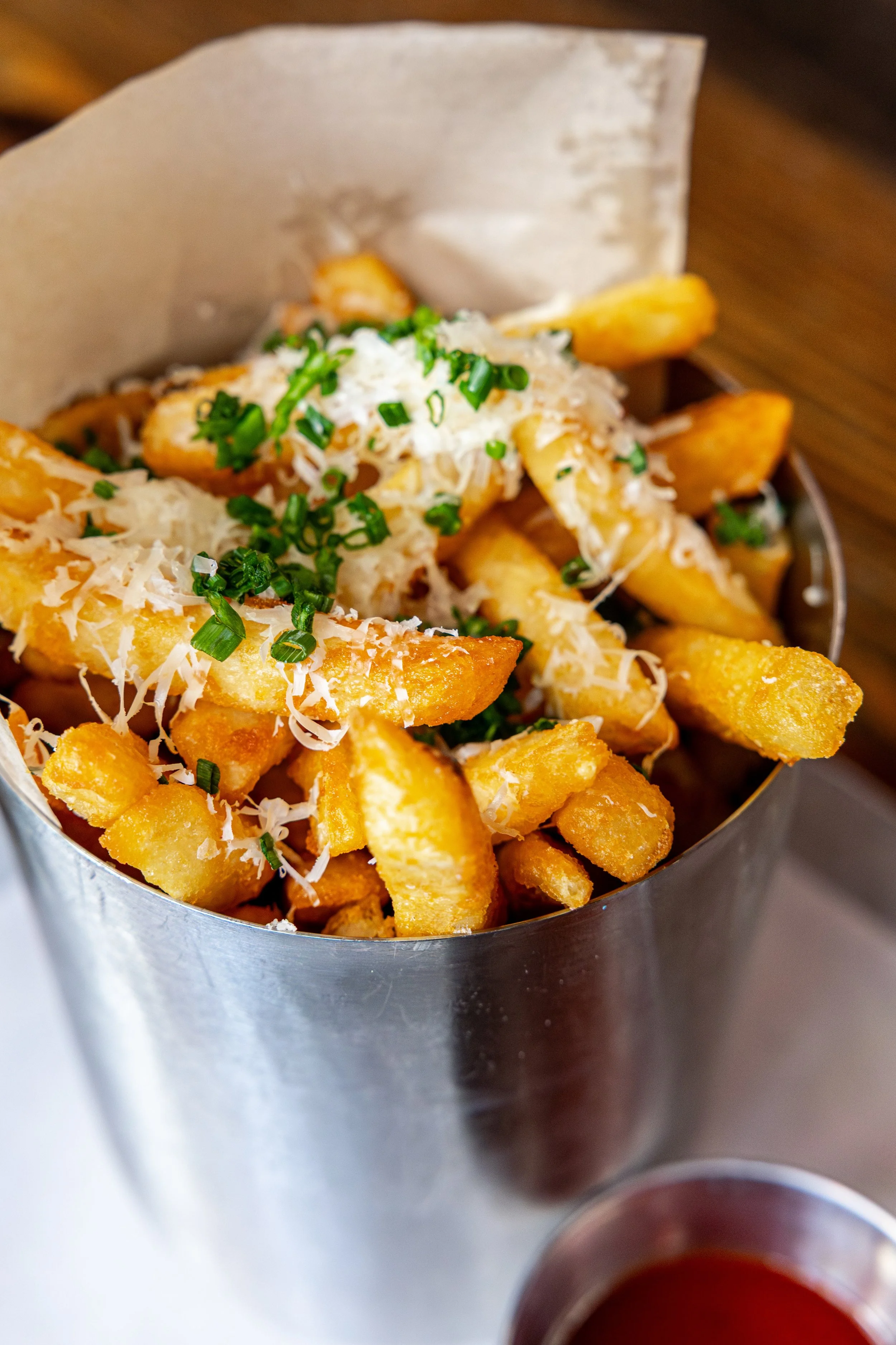 Fried potato wedges topped with grated cheese and chopped green onions in a metal serving container, with a small cup of dipping sauce nearby.
