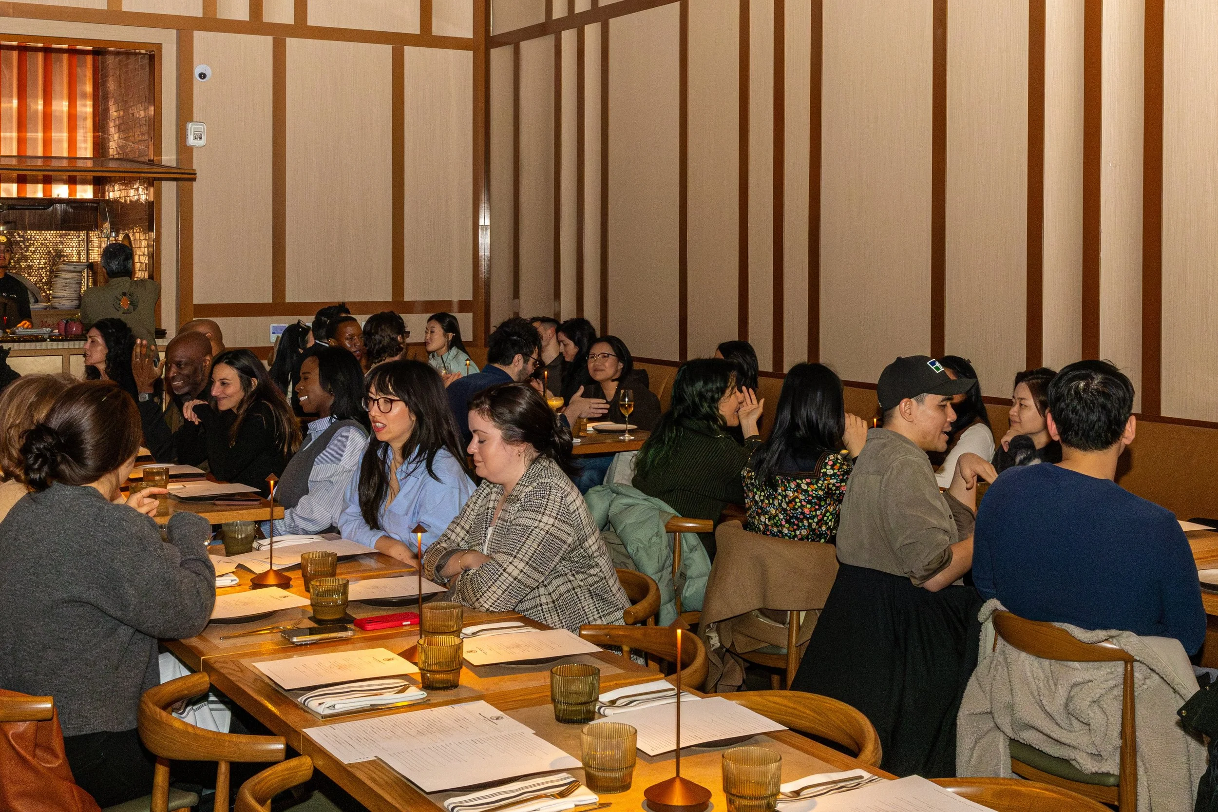 A diverse group of people sitting at tables in a restaurant or event space, engaged in conversation and enjoying drinks, with some looking towards the front and others talking among themselves.