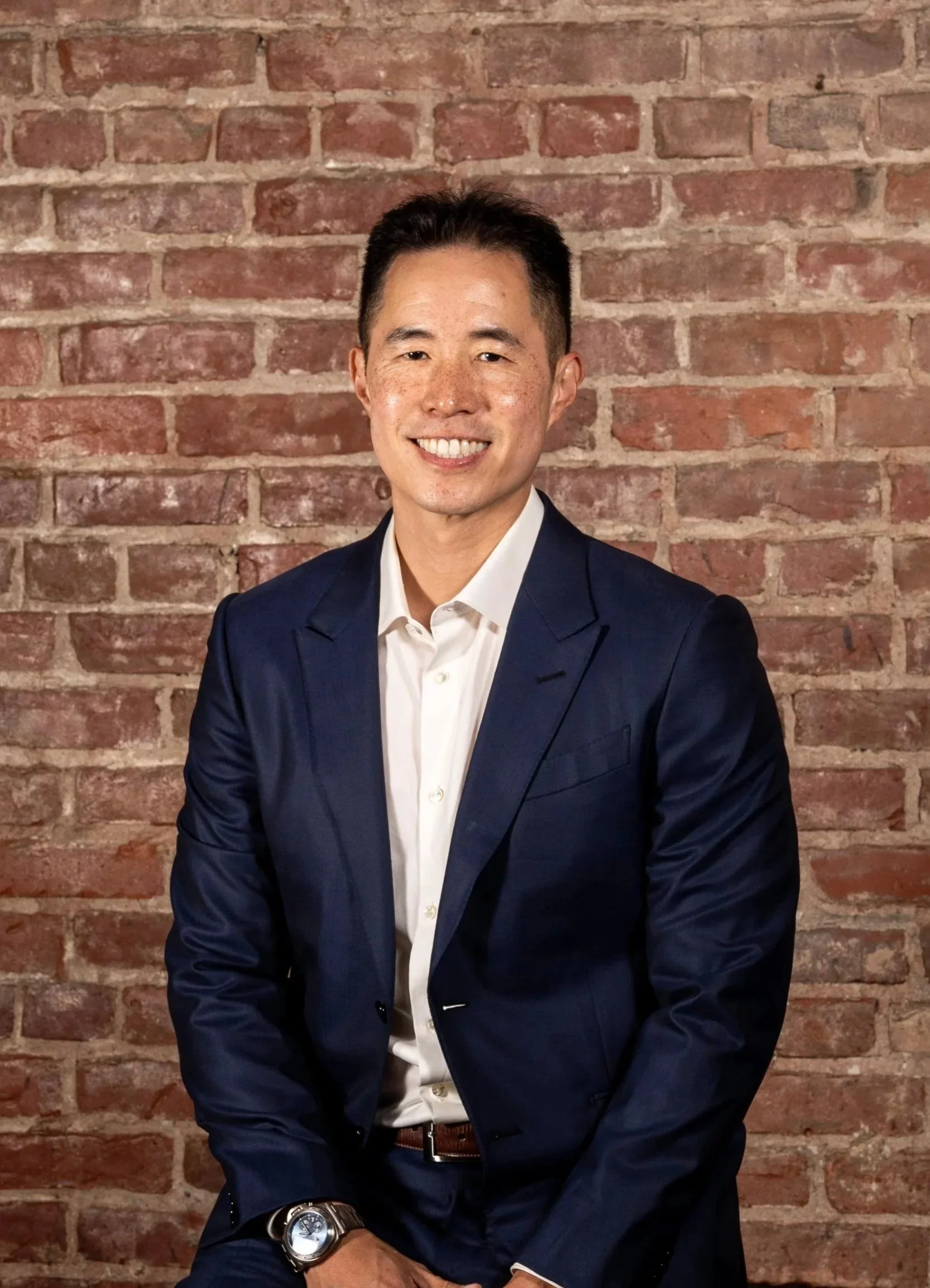 A man in a navy blue suit with a white shirt, smiling, sitting in front of a brick wall. He has short black hair, freckles, and is wearing a large watch on his left wrist.