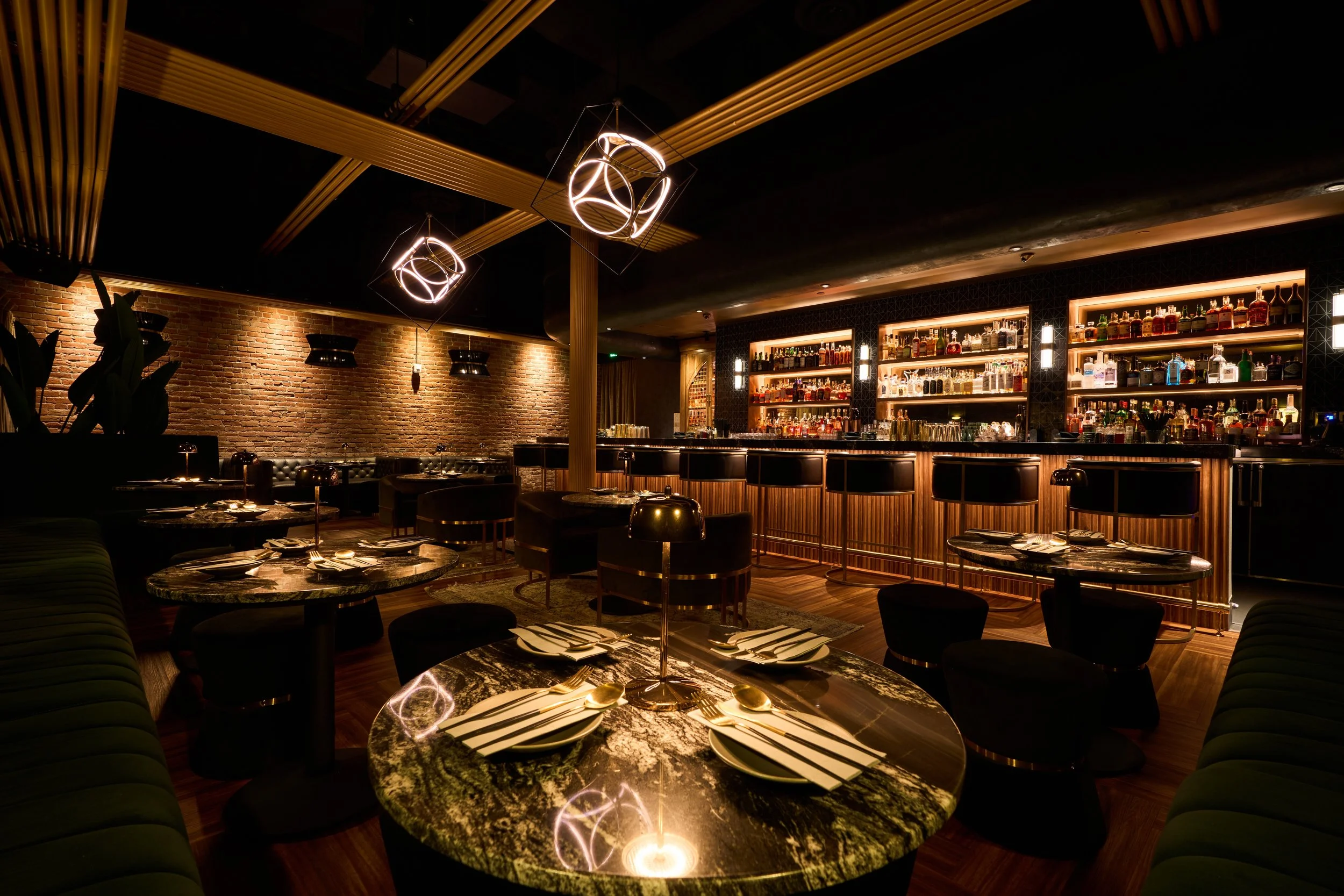 An upscale bar and restaurant interior with illuminated shelves of liquor bottles, decorative lighting fixtures, brick walls, and elegant tables set for dining.