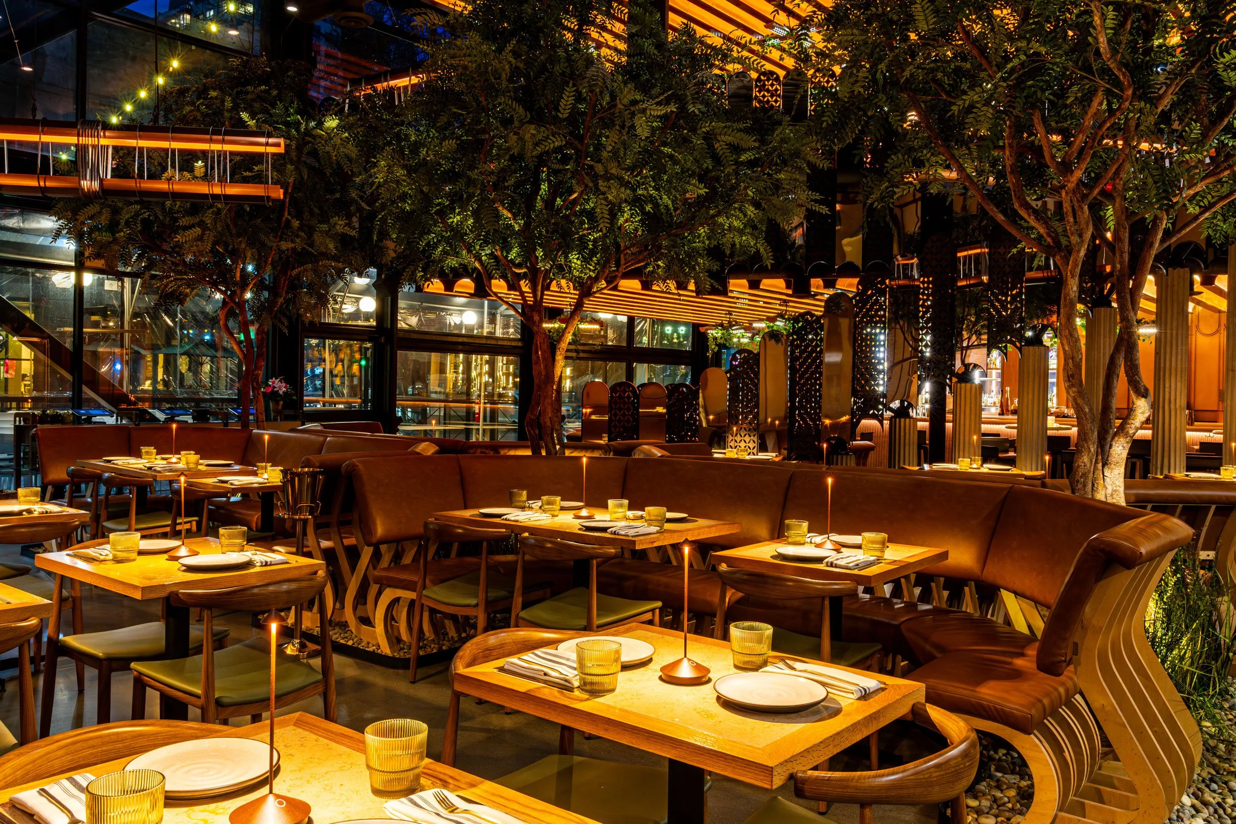 Interior of a modern restaurant with tables set for dining, featuring warm lighting, lush greenery including trees, and decorative partitions with lighting accents.