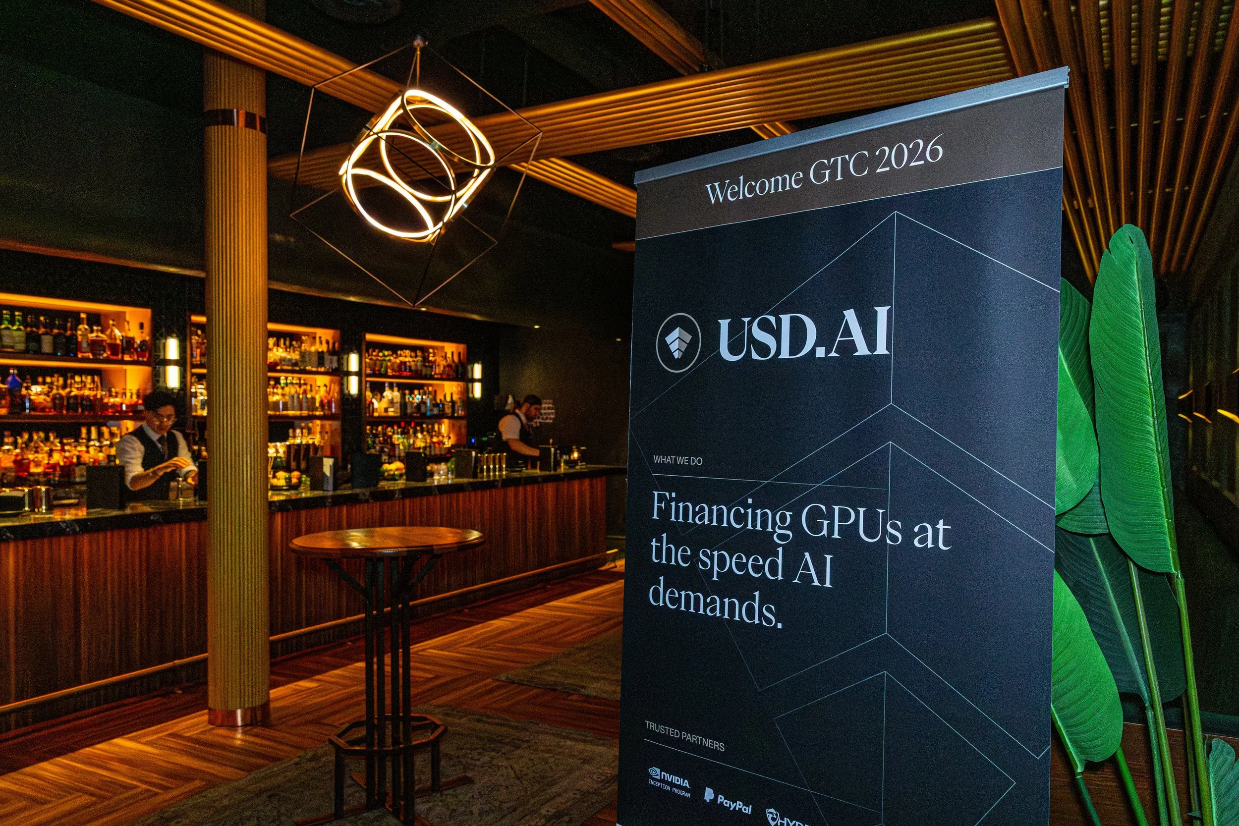 A bar scene at a conference showing a large sign with the text "Welcome GTC 2026" and "USD.AI". The bar has bottles of alcohol on illuminated shelves with bartenders serving drinks. Modern lighting fixtures hang from the ceiling, and a small round ta