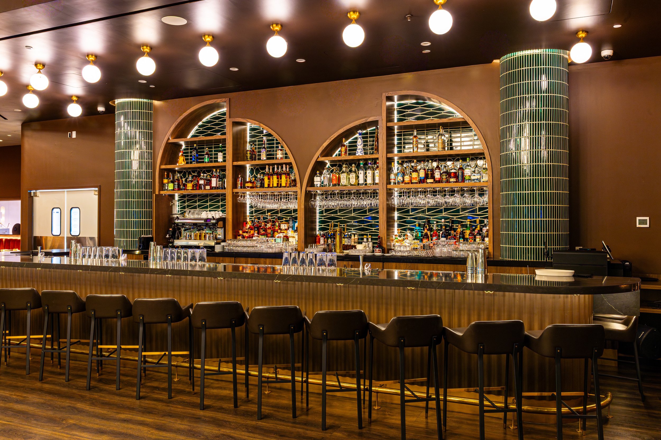 Inside view of a bar area with a curved wooden countertop, black chairs, and a backlit wooden shelving displaying various bottles of liquor and glassware. There are round white ceiling lights and two large green tiled columns.