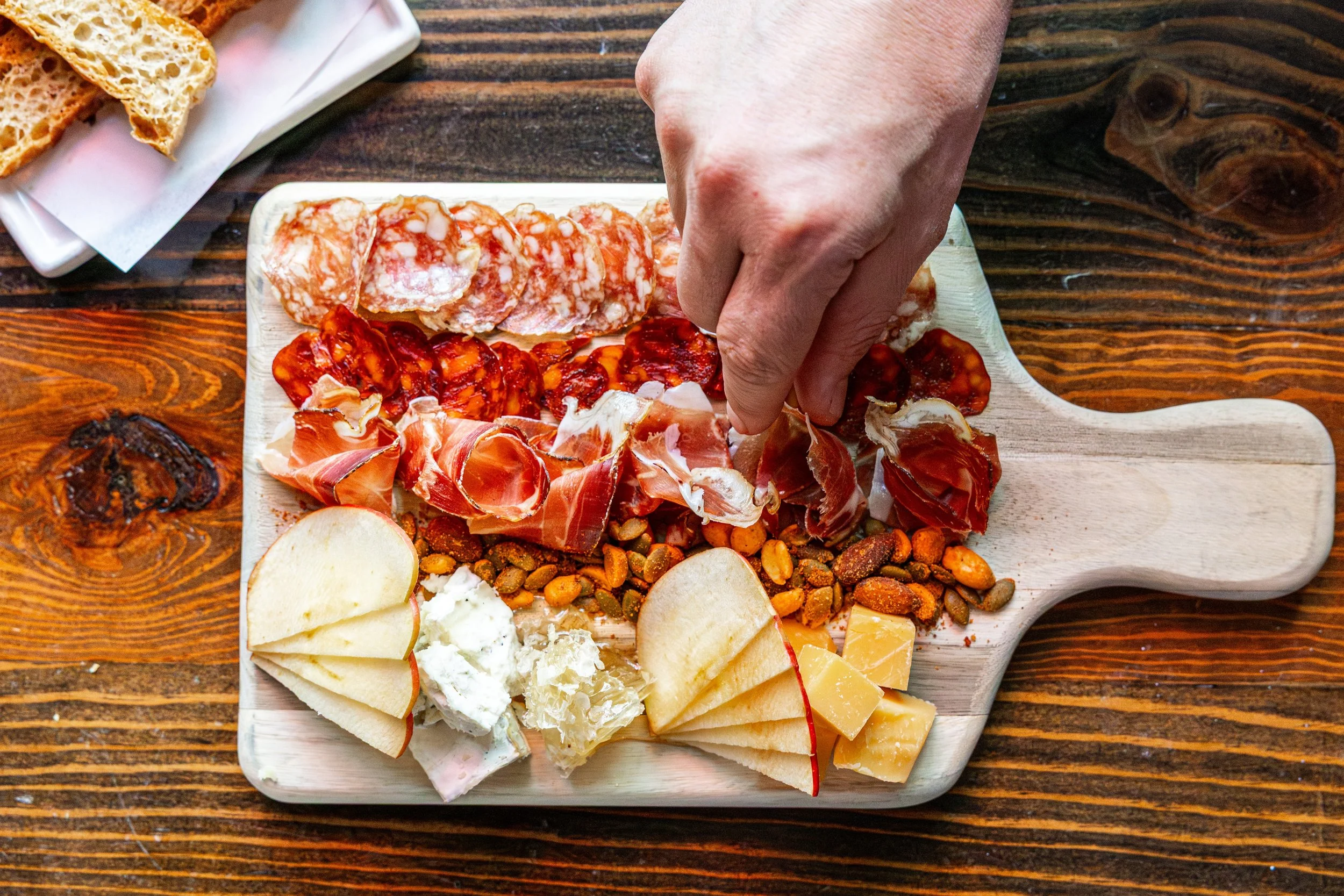 A wooden cheese and charcuterie board with sliced cheeses, sliced apples, prosciutto, roasted nuts, and various cured meats placed on a rustic wooden table.