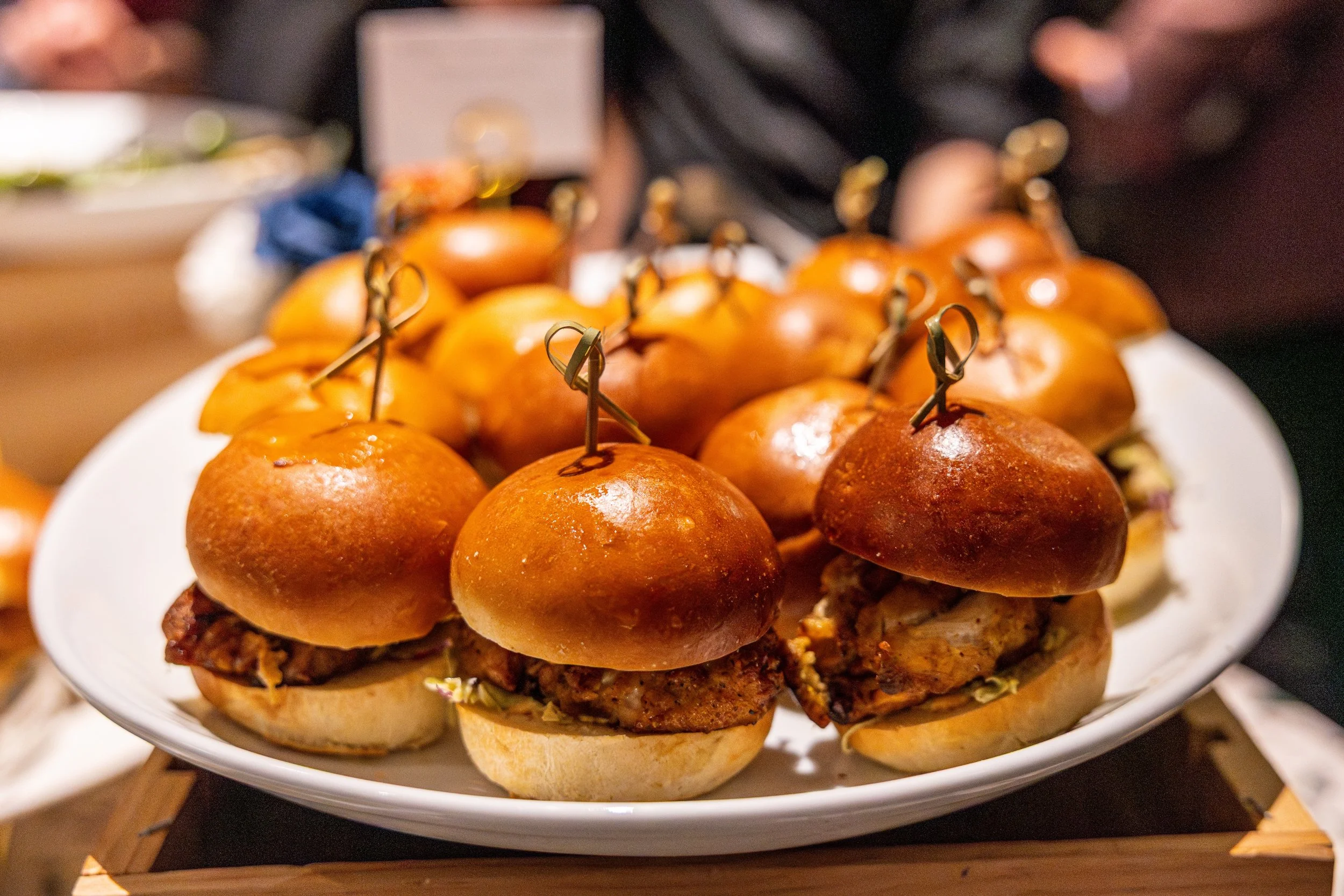 Plate of small slider sandwiches with roasted turkey or chicken, topped with mini sandwich buns, and skewered with small metal picks. The background has a blurred setting with other dishes and a person's arm visible.