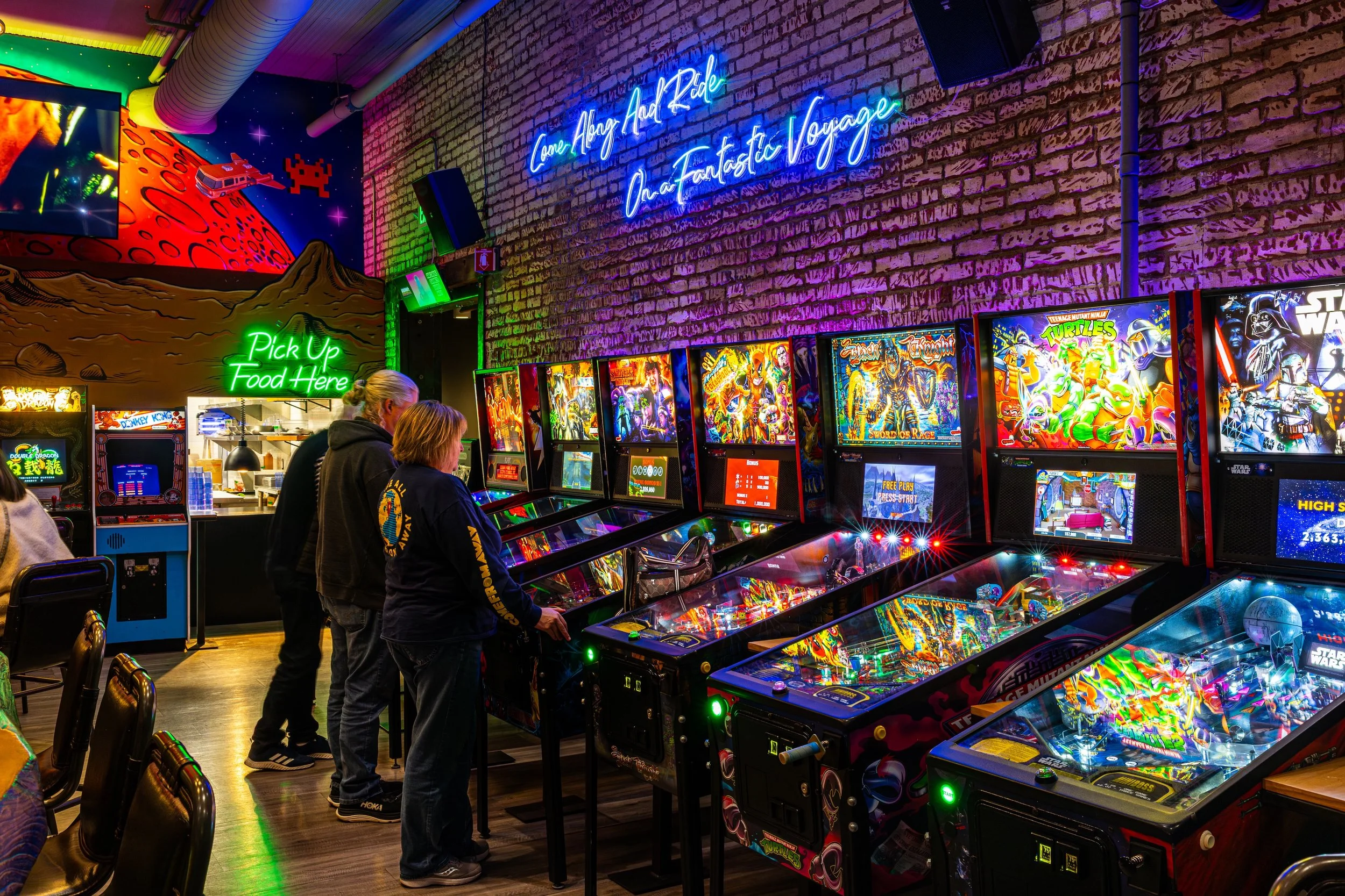 People playing pinball machines inside an arcade with colorful neon signs and space-themed wall art.