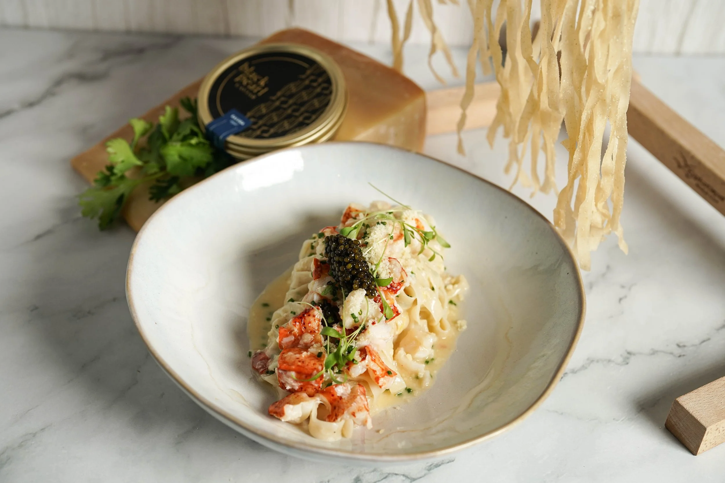 Lobster and caviar pasta dish in a white bowl on a marble surface with a wooden board and stone in the background.