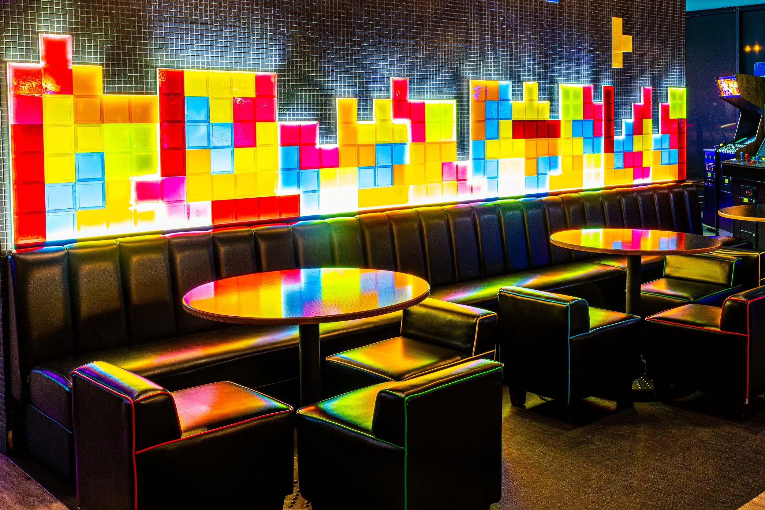 Colorful illuminated Tetris-style block art on a dark wall with black leather seating and tables in a lounge or bar setting.