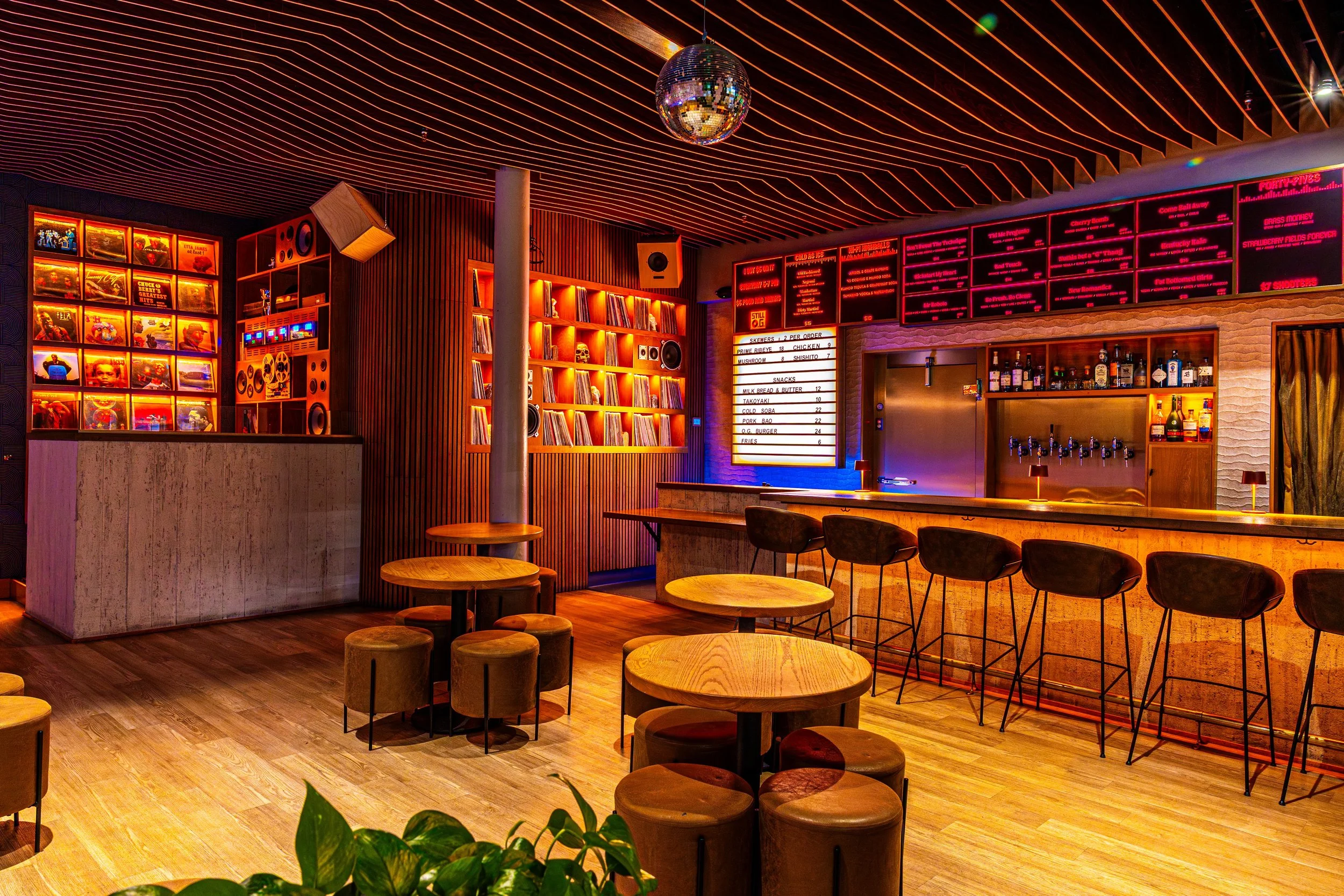 Interior of a bar or music venue with wooden tables and chairs, shelves of records, red and orange neon lighting, a disco ball on the ceiling, and a digital menu board.