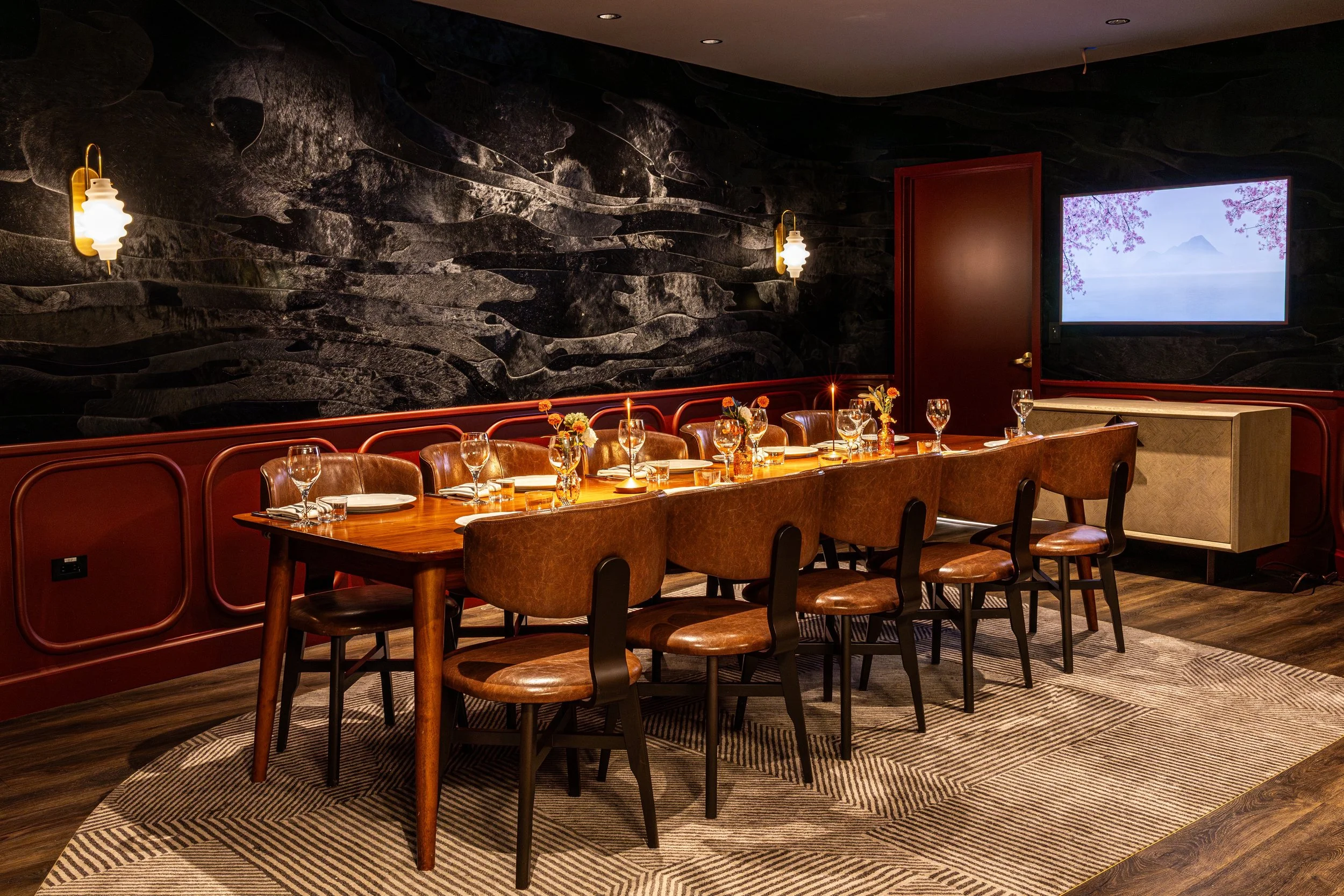 A cozy restaurant dining area with a long wooden table set with wine glasses, small flower arrangements, and candles. The room has a dark, textured wall with two wall-mounted lights, and a large window showing pink flowering trees and a mountain in t