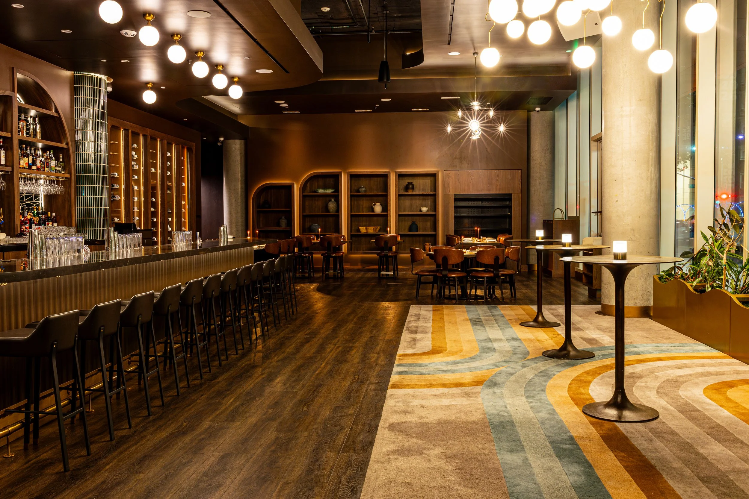 An upscale restaurant interior with a bar on the left, rows of barstools, and wooden shelving on the back wall with decorative items. The right side features round tables with chairs, large windows, and modern lighting fixtures hanging from the ceili
