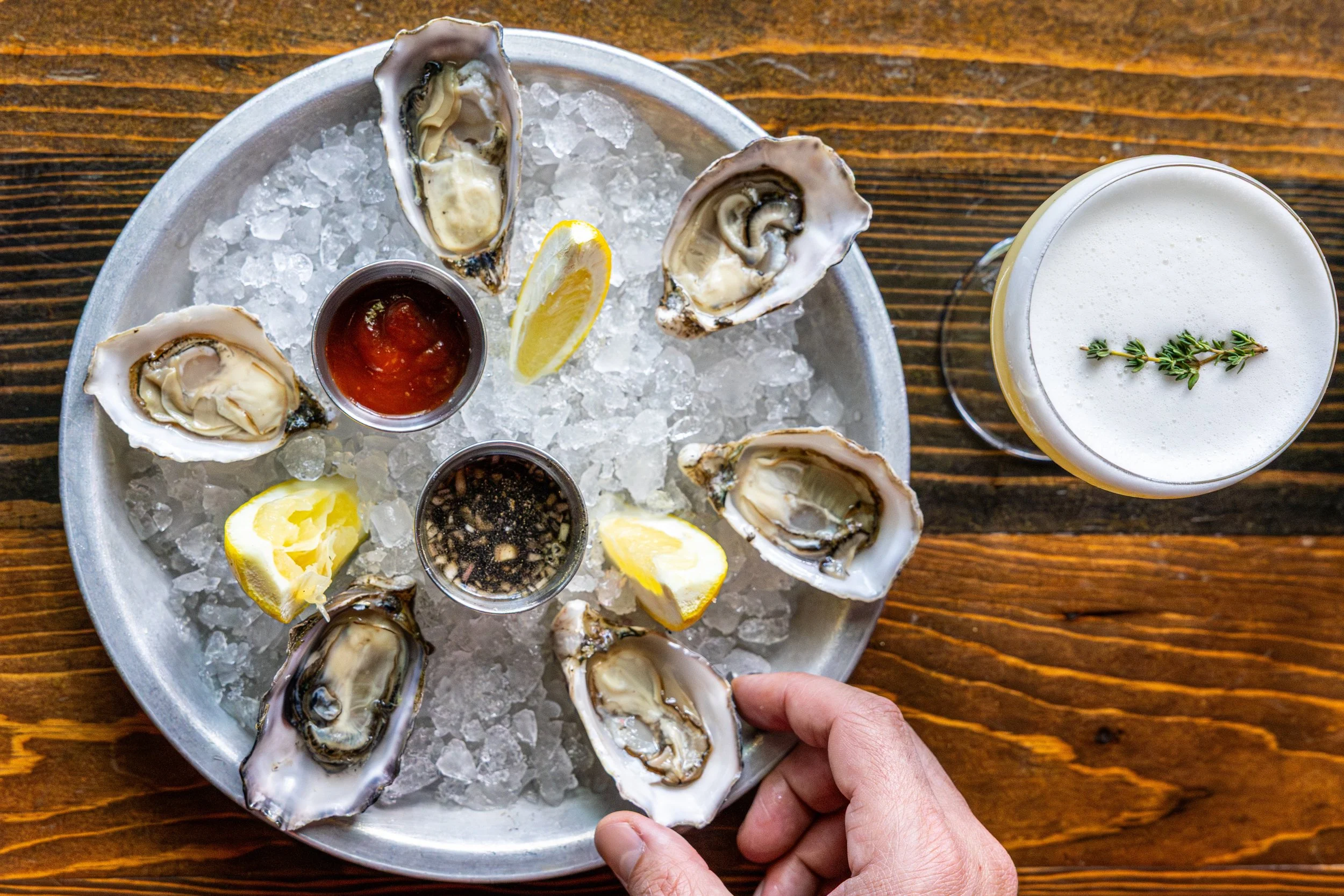 A metal tray filled with ice and fresh oysters on the half shell, garnished with lemon wedges and accompanied by dipping sauces, with a glass of creamy beverage garnished with a sprig of thyme next to the tray on a wooden surface.