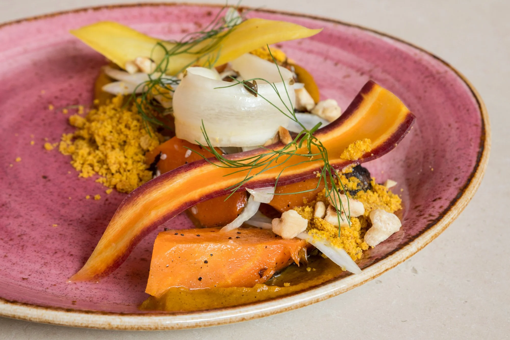 Close-up of a pink ceramic plate with a gourmet dish featuring roasted butternut squash, a dollop of cream, crumbled nuts, yellow crumble, and garnished with fresh herbs.