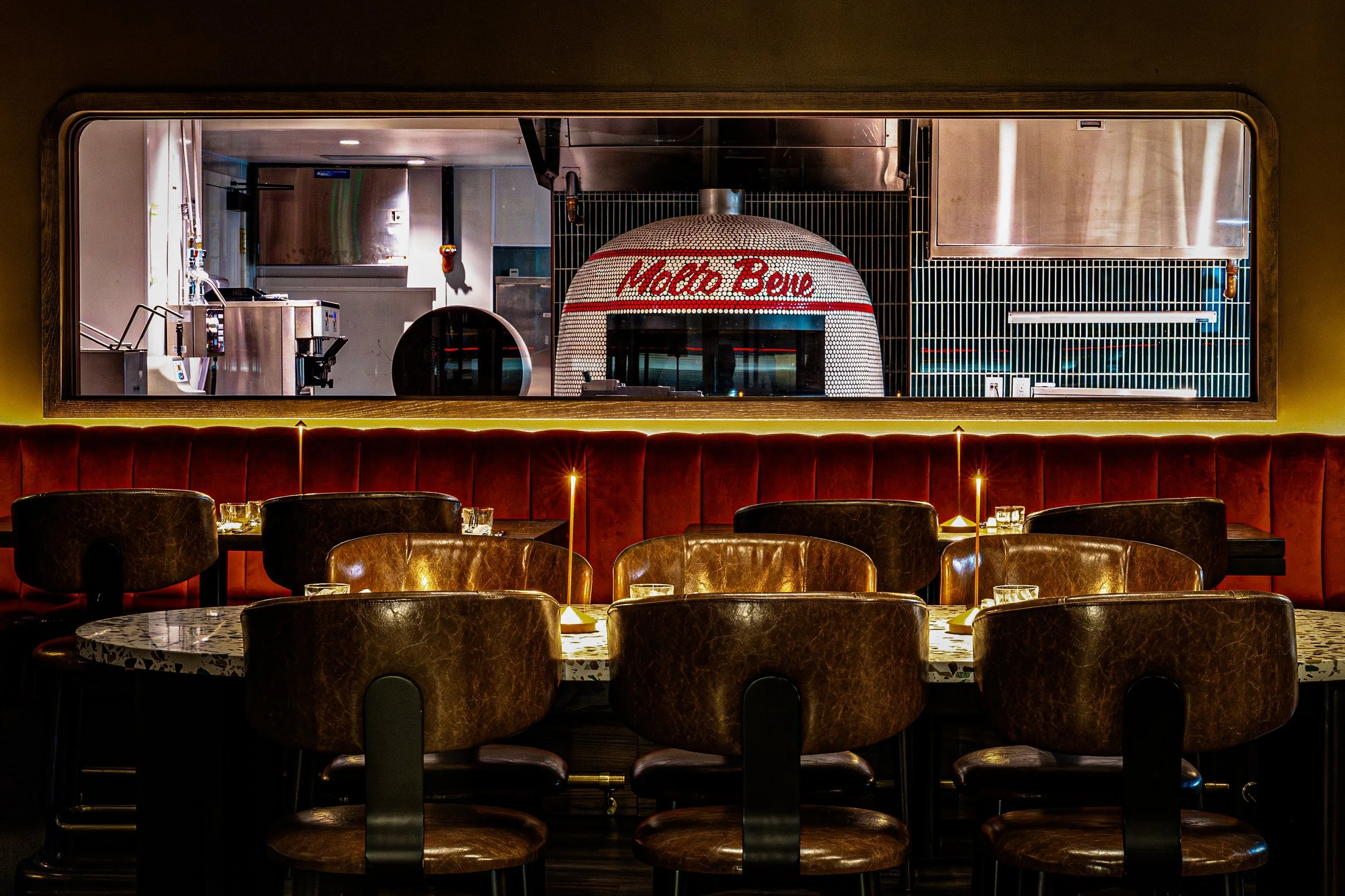 A view through a window of a kitchen with a pizza oven that has 'Made Bear' written in red on the dome, with tables and chairs in a restaurant setting in the foreground.