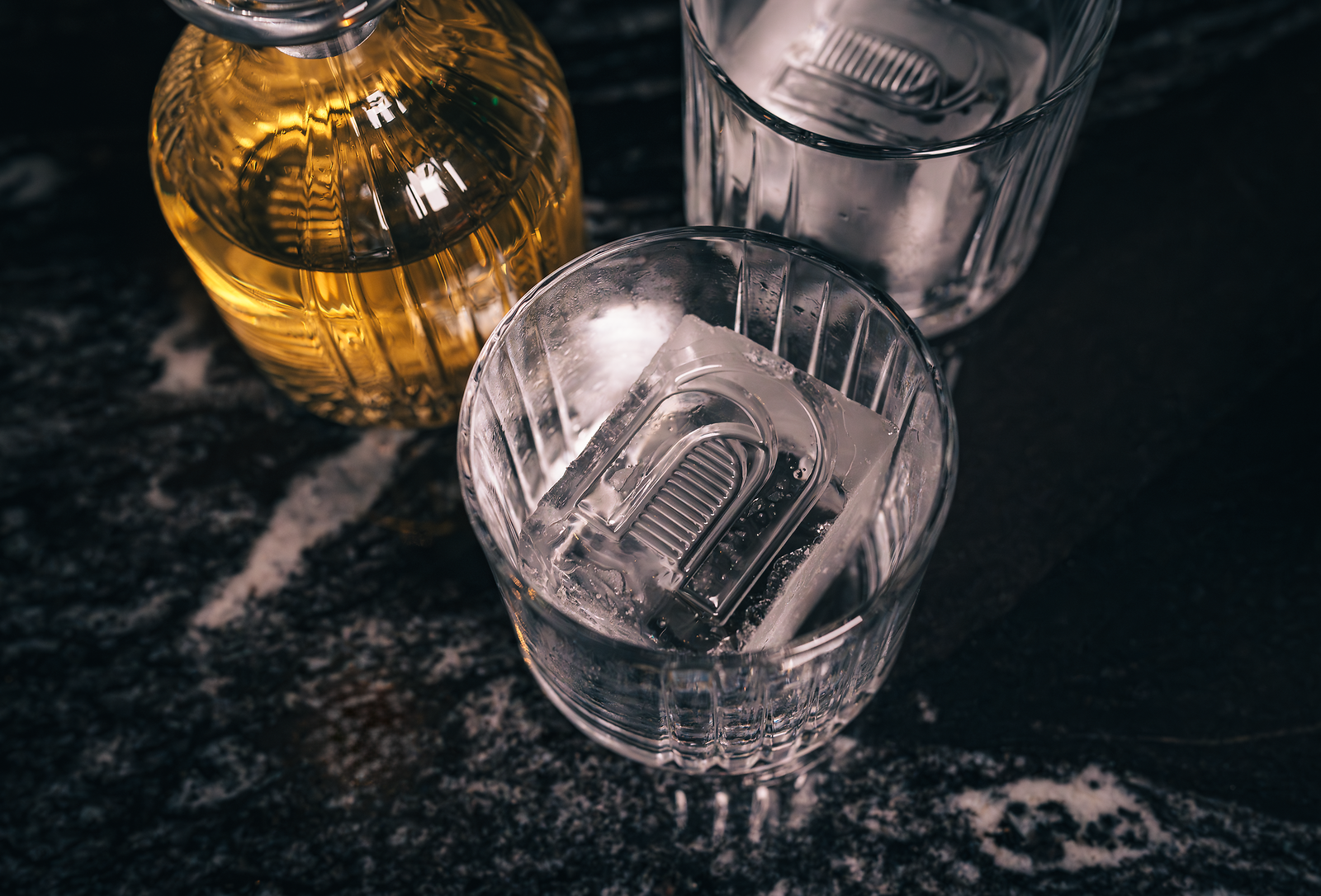Close-up of a glass with ice cubes and metal bar inside, a bottle of whiskey, and a glass of clear liquid with ice and metal bar, on a dark marble surface.