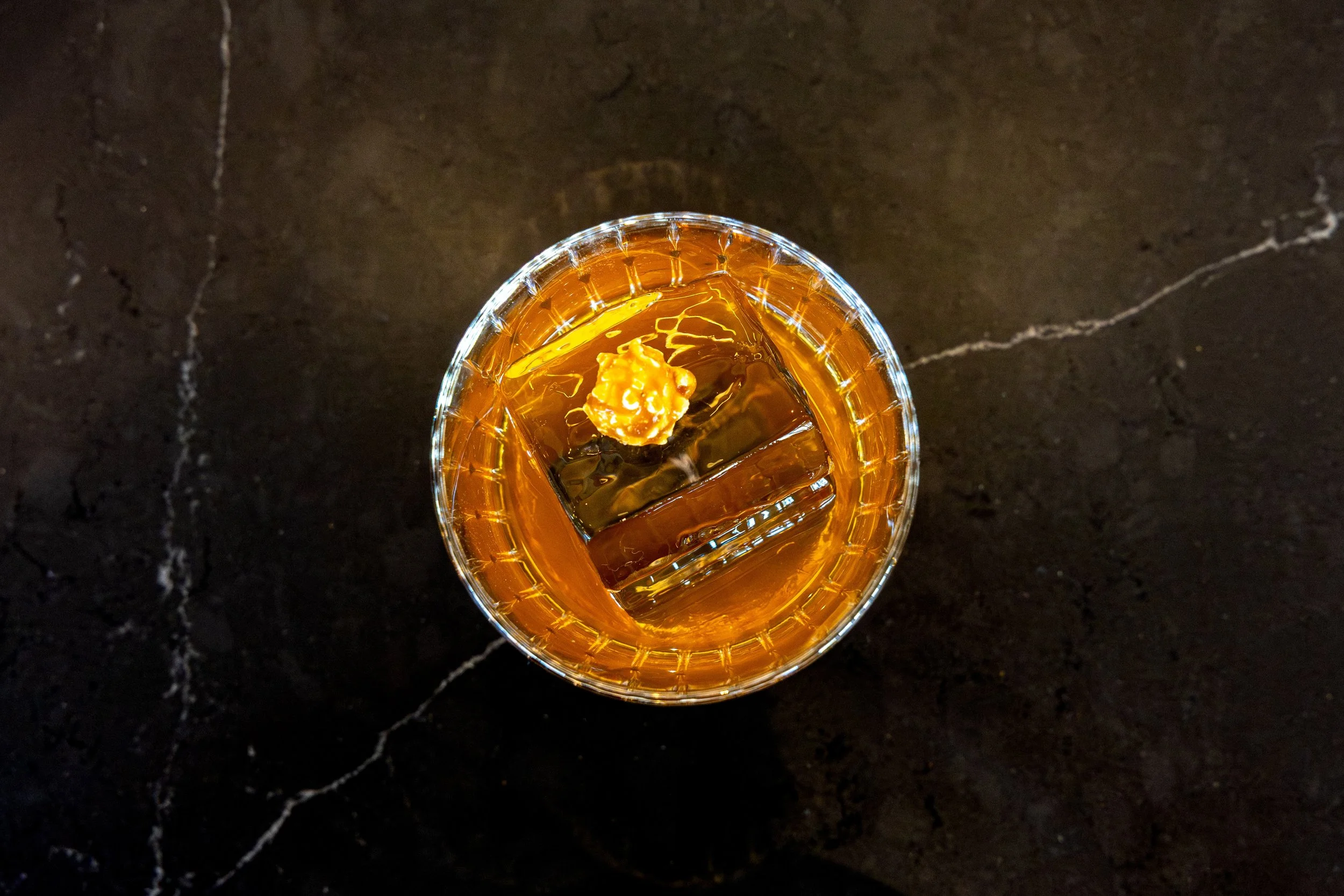 A glass of iced whiskey with a lemon twist on a dark marble surface.