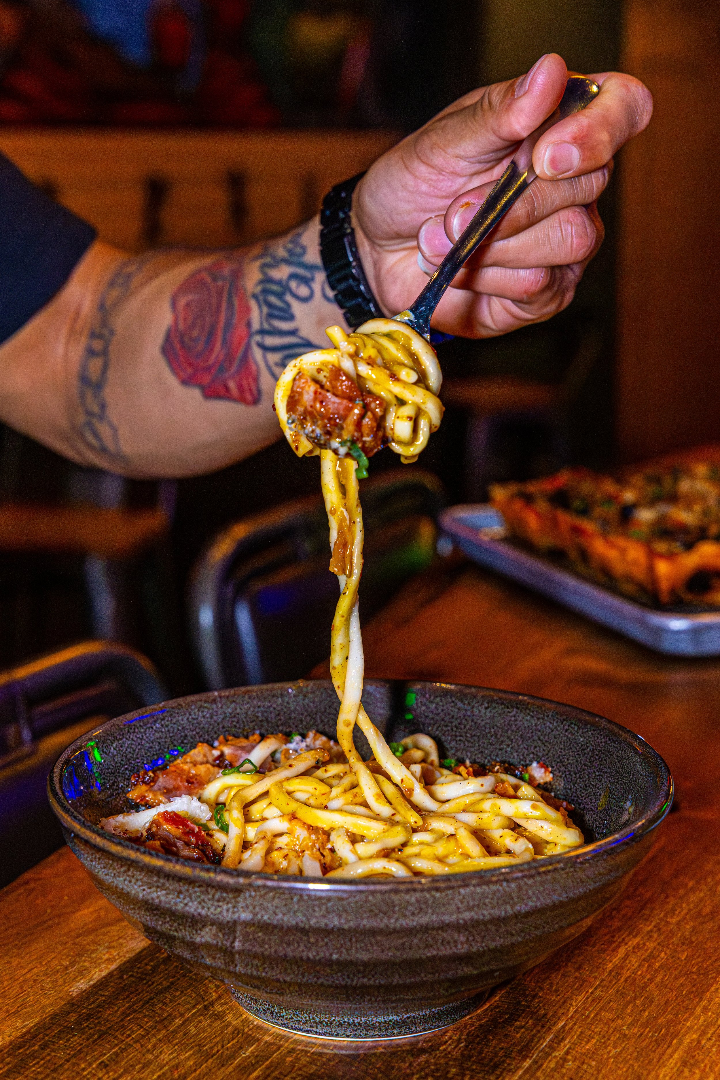 Person with tattooed arm holding fork with noodles and sauce, about to eat from a bowl of Asian-style noodles on a wooden table.