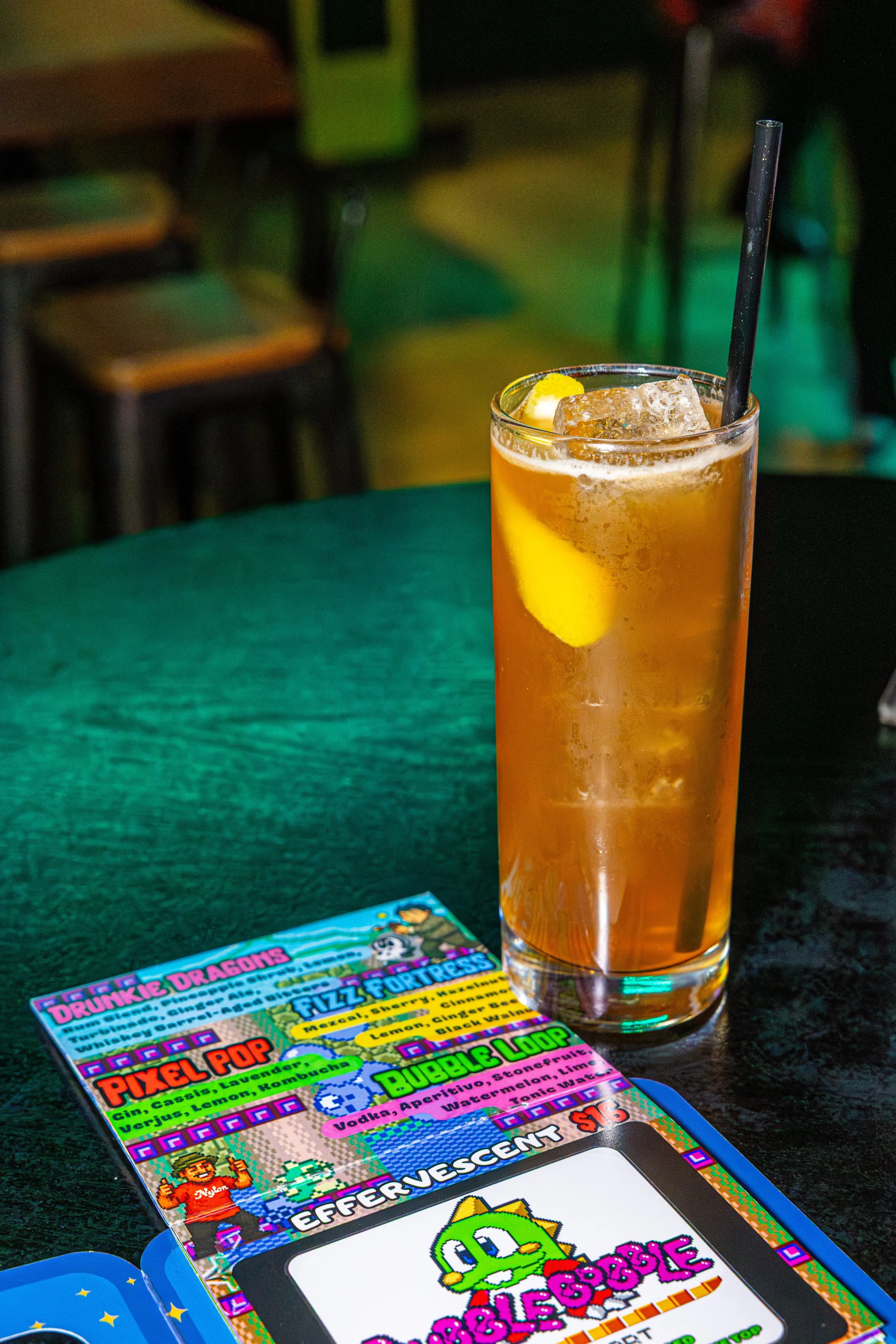 A tall glass of iced tea with a lemon wedge and a black straw, on a dark table at a bar or restaurant. Partially visible colorful game magazine or brochure with pixel art graphics on the table.