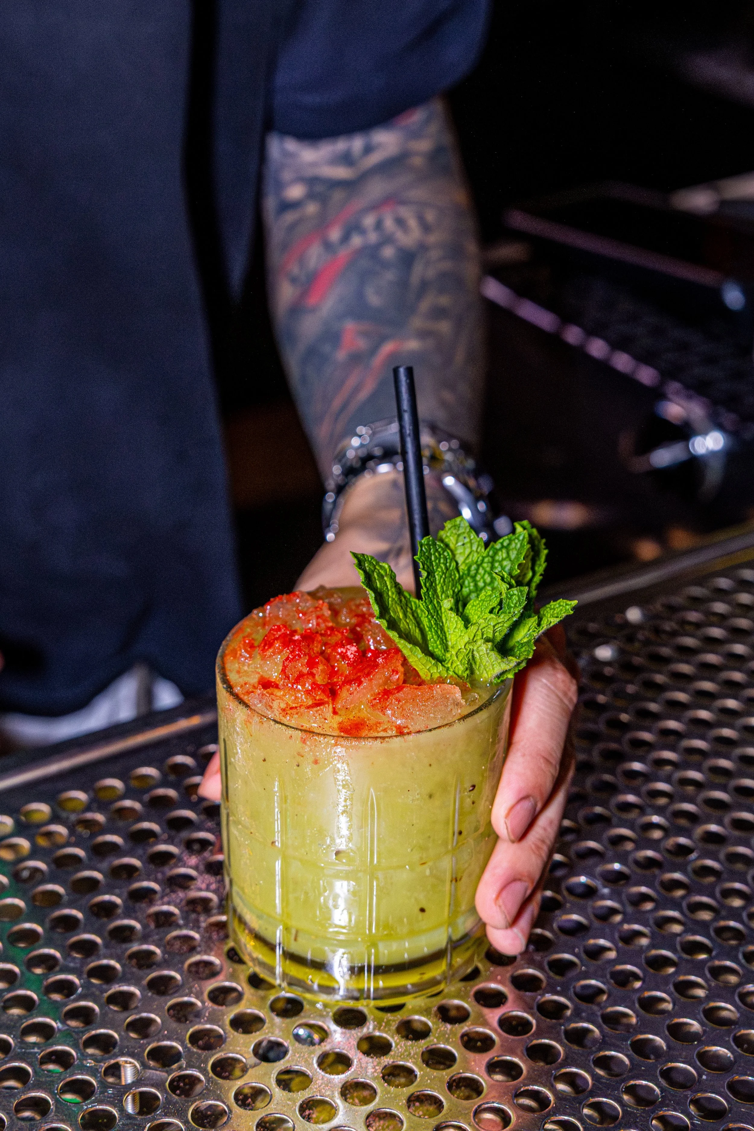 A hand holding a yellow cocktail garnished with mint leaves and red chili flakes, in front of a person with a tattooed arm wearing a dark shirt, sitting at a metal table.