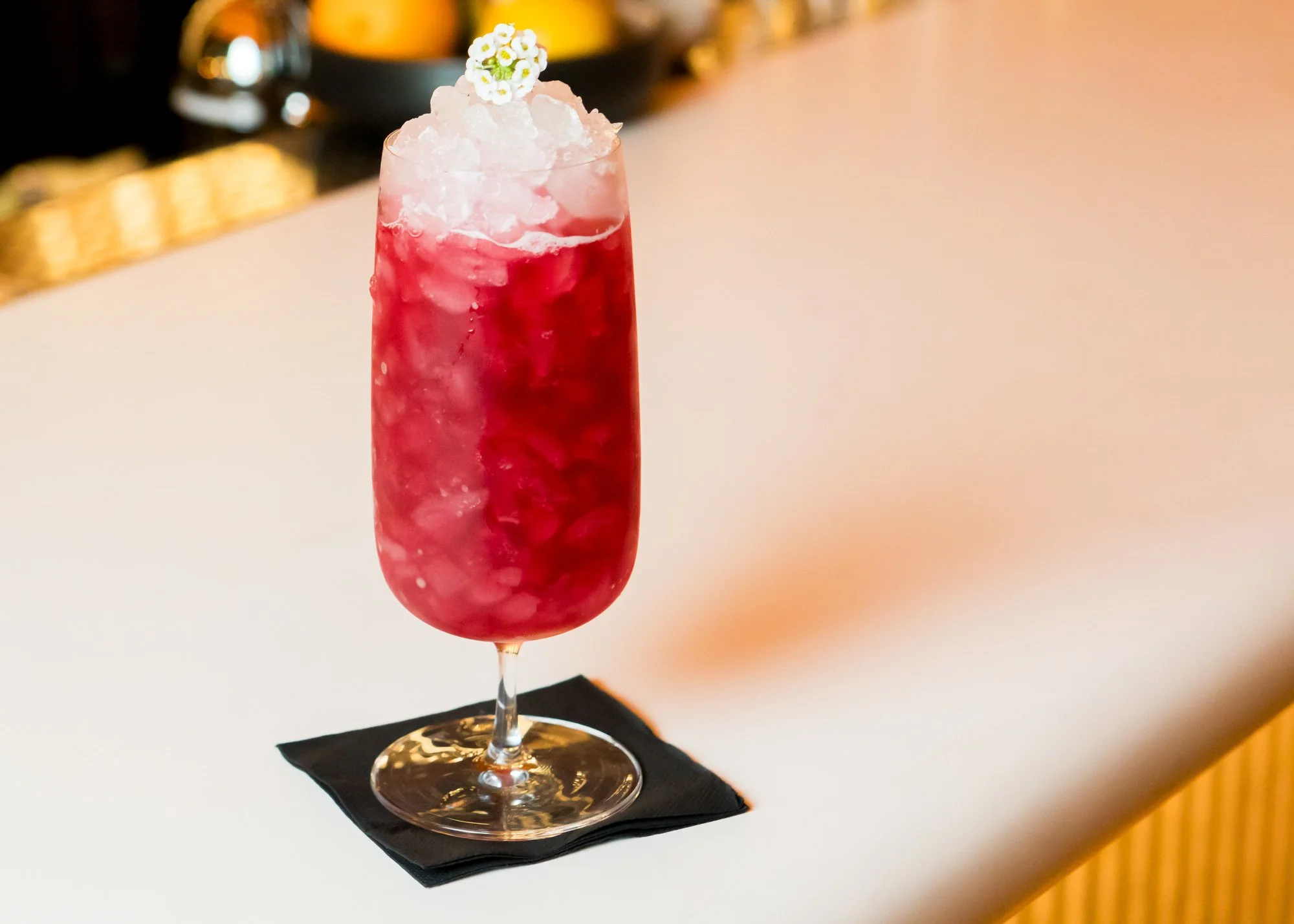 A tall glass filled with a red, crushed ice beverage topped with a small white flower. The drink is placed on a black napkin on a bar counter with a blurred background.