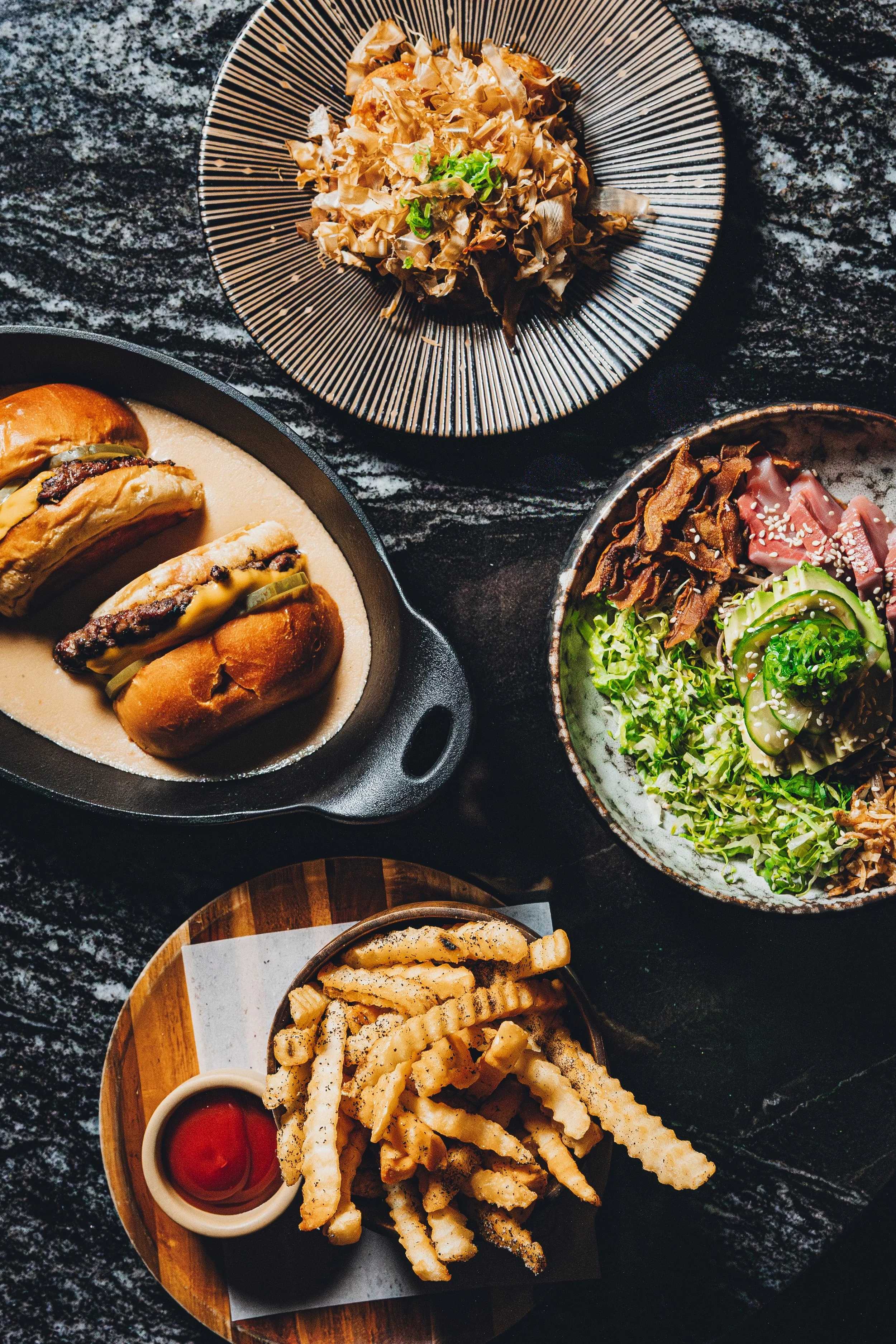 Various dishes of Japanese-inspired food, including a plate of fried chicken and fries with ketchup, a bowl of mixed greens and sliced meat with sesame seeds, a bowl of fried cheese sticks with ketchup, and small dishes of sauces, all on dark surface
