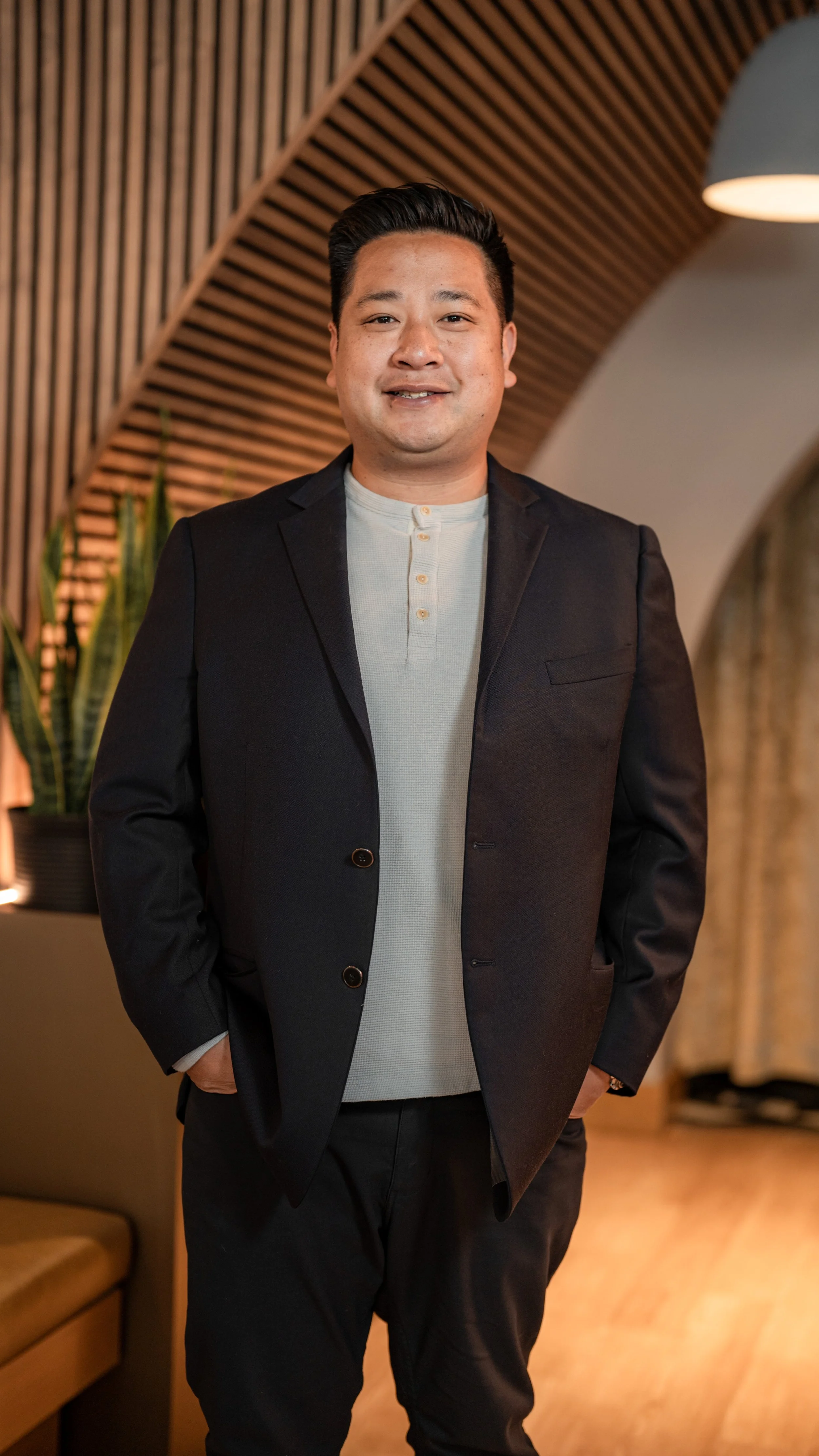 A man wearing a light gray henley shirt and a black blazer standing indoors with a wooden wall and plants in the background.