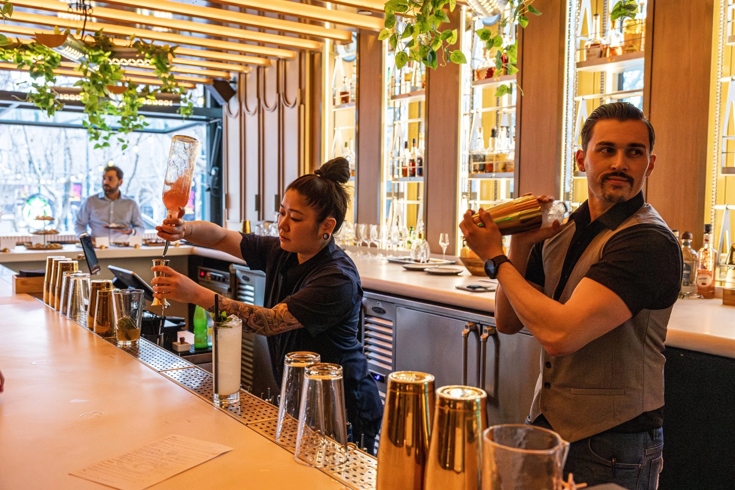 Bartenders are preparing drinks at a modern bar with gold accents, tall glassware, and a well-lit, decorative background. One bartender is pouring a drink from a large bottle, while another looks to the side holding a shaker.