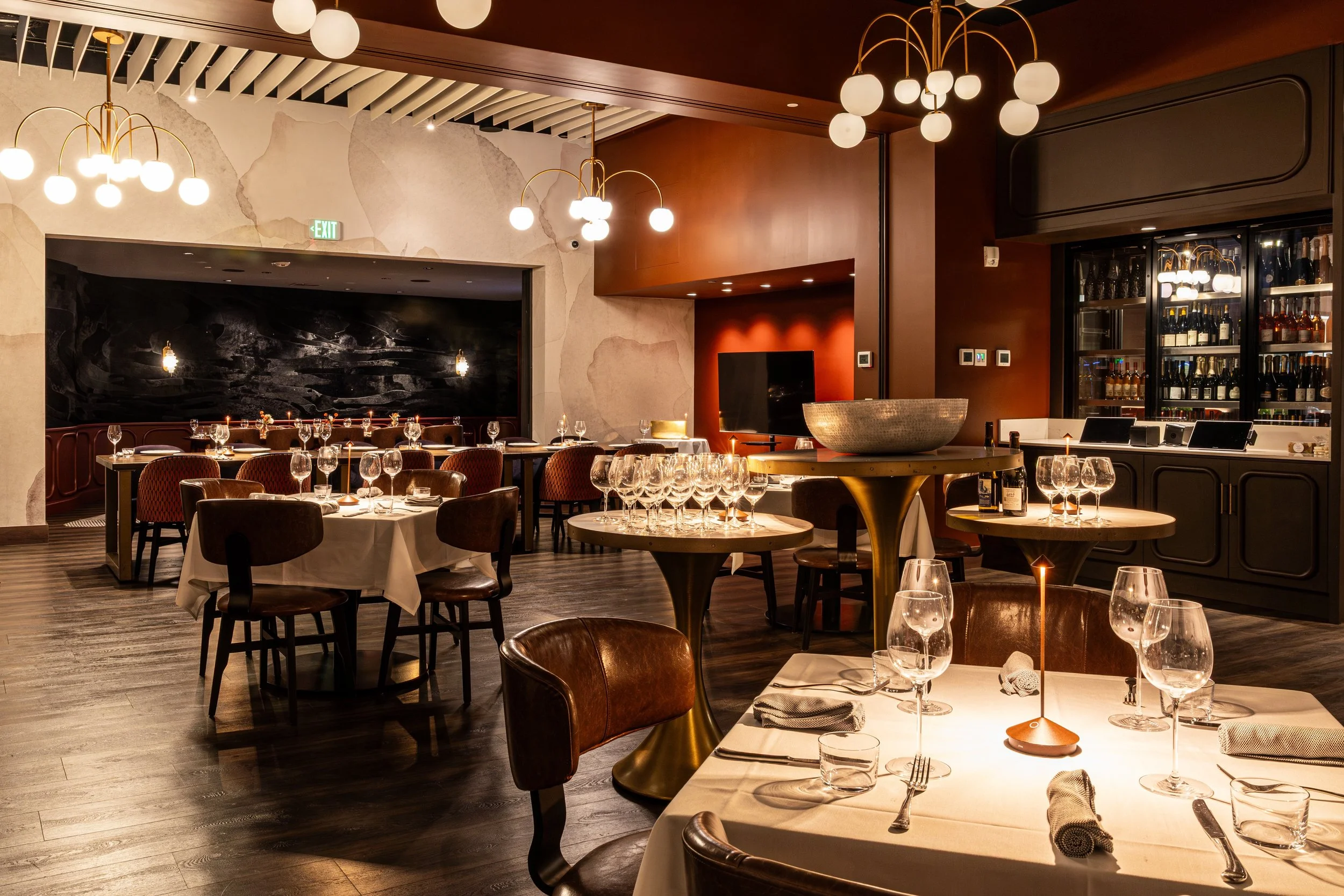 Elegant restaurant dining area with round tables, wine glasses, and soft lighting, featuring modern light fixtures and wine display cabinets.