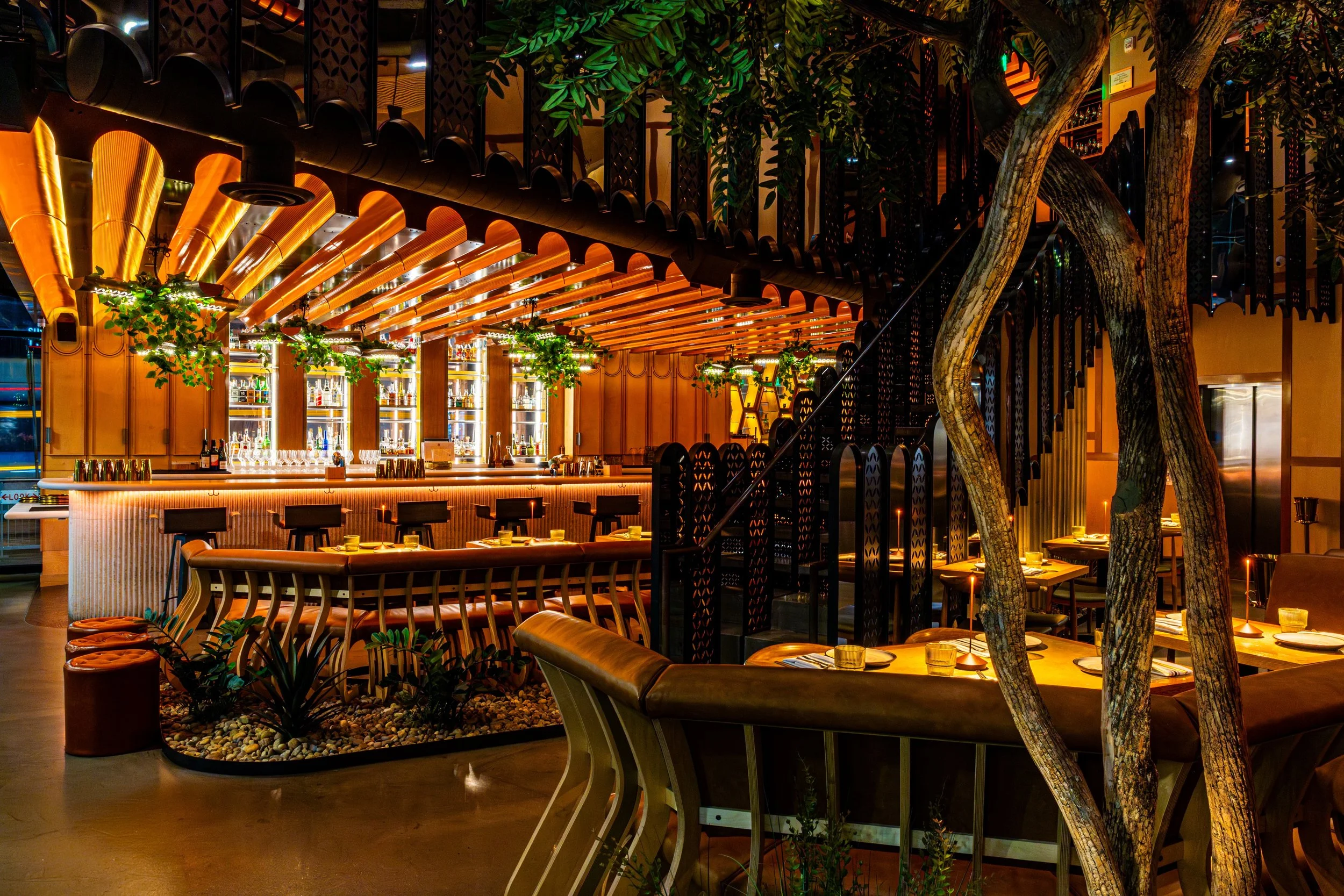 A modern restaurant interior with a bar area illuminated by warm lighting, featuring wooden accents, lush green plants, and large trees integrated into the decor.