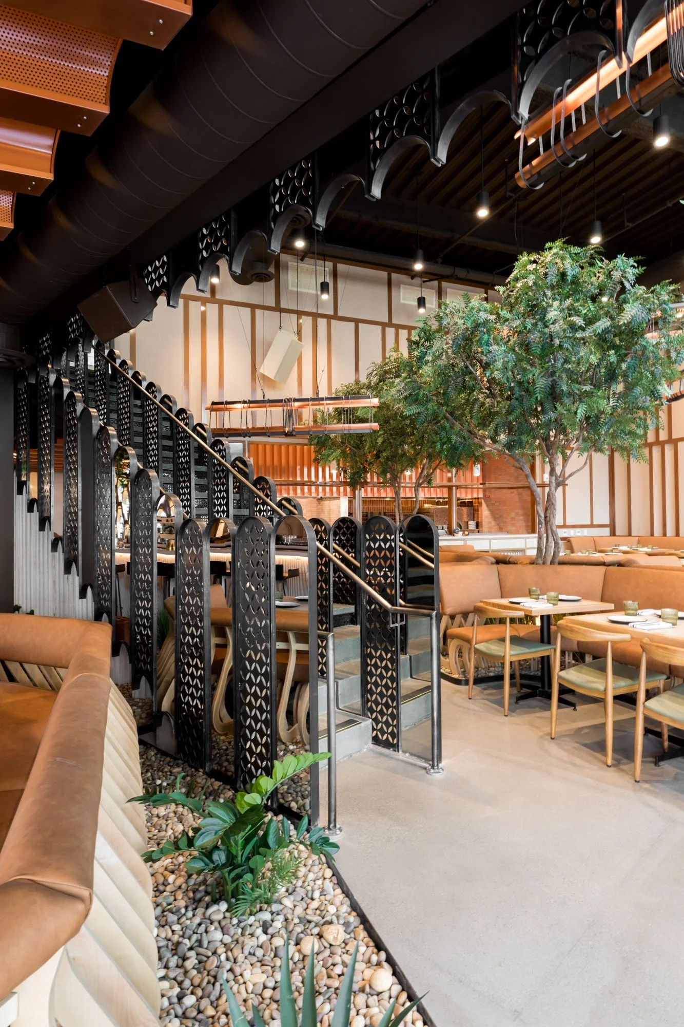 Interior of a modern restaurant with a large potted tree, beige seating, black metal decor, and warm lighting.
