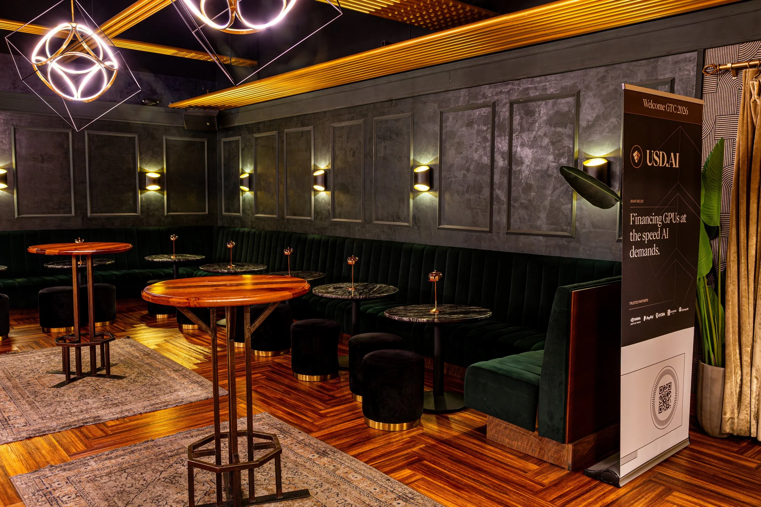 Interior of a modern lounge area with dark walls, green velvet seating, multiple tables, and unique hanging and wall lighting fixtures. There is a welcome sign and a QR code stand near the entrance.