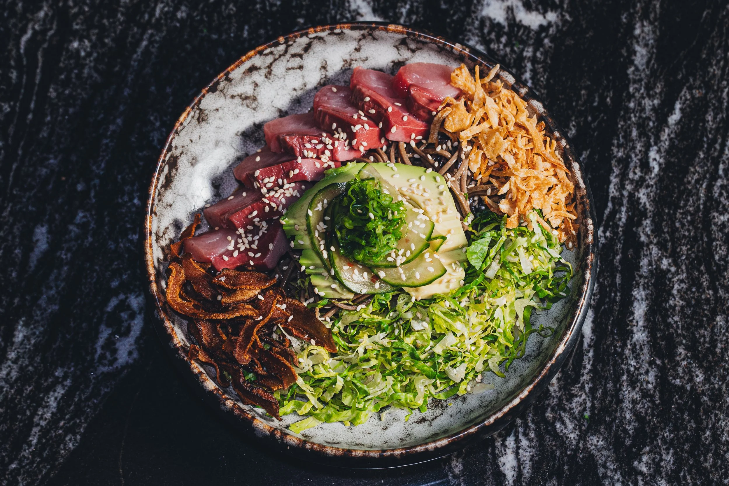 A bowl containing slices of raw beef, cucumber, shredded cabbage, shredded carrots, and crispy fried onions, garnished with sesame seeds, placed on a dark marble surface.