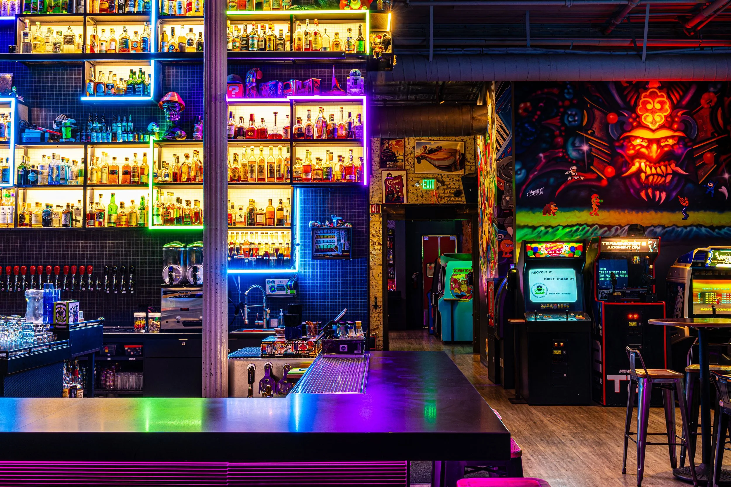 A vibrant indoor arcade with a brightly lit bar on the left, showcasing shelves of alcohol bottles illuminated by colorful neon lights, and arcade game machines on the right with a colorful mural on the wall featuring a fierce, stylized face with glowing eyes.
