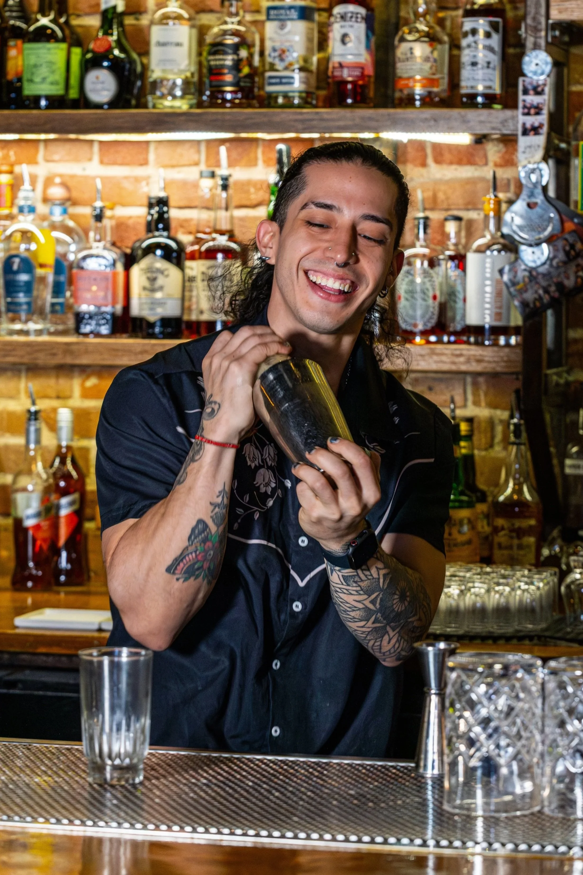 A smiling bartender with tattoos and piercings holding a cocktail shaker behind a bar with various bottles of alcohol on shelves.