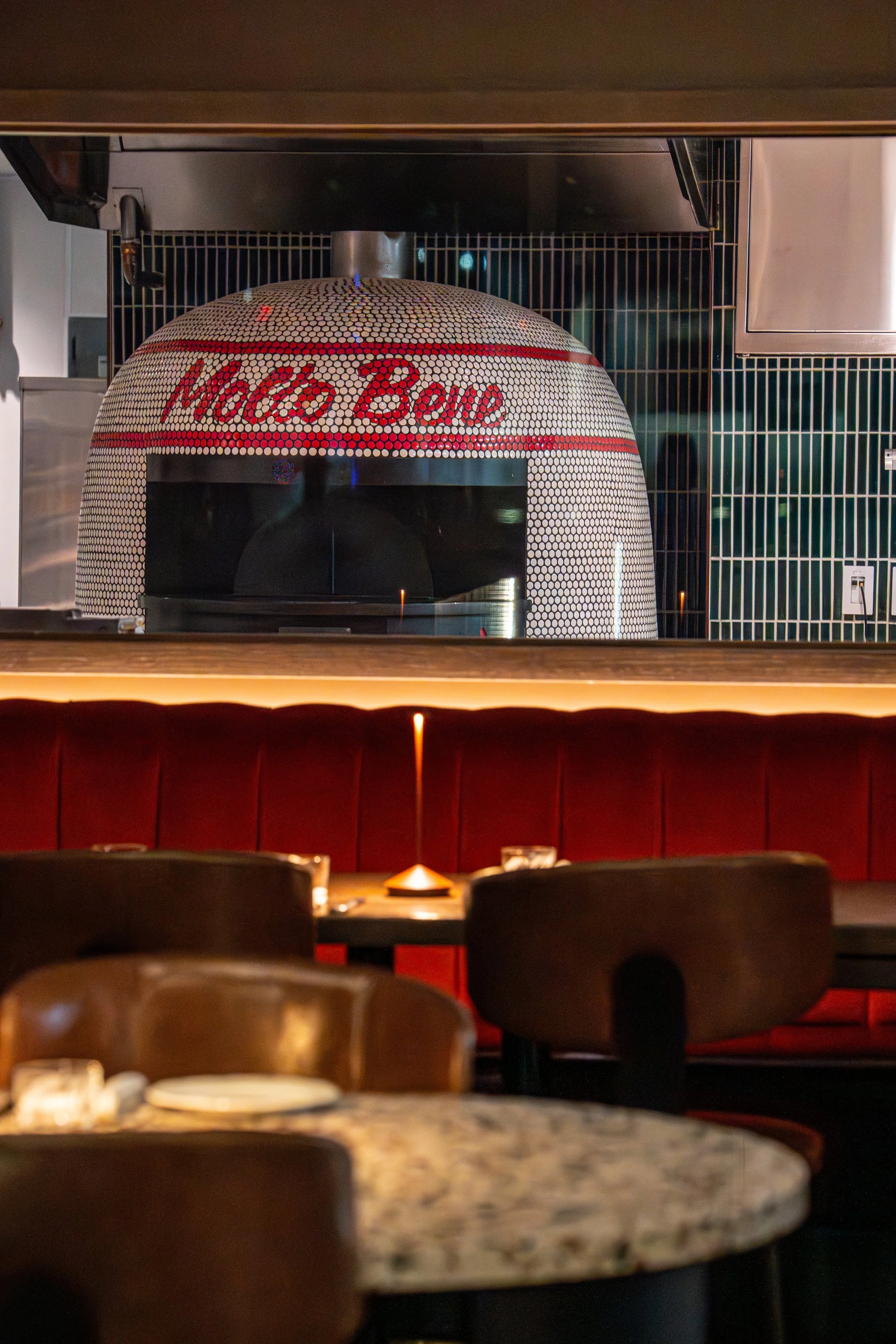 View of a pizza oven with a domed tile exterior featuring the name 'Macho Bow' written in red, situated in a restaurant kitchen. The oven is surrounded by black metal grids and is playing a central role in the kitchen setting.